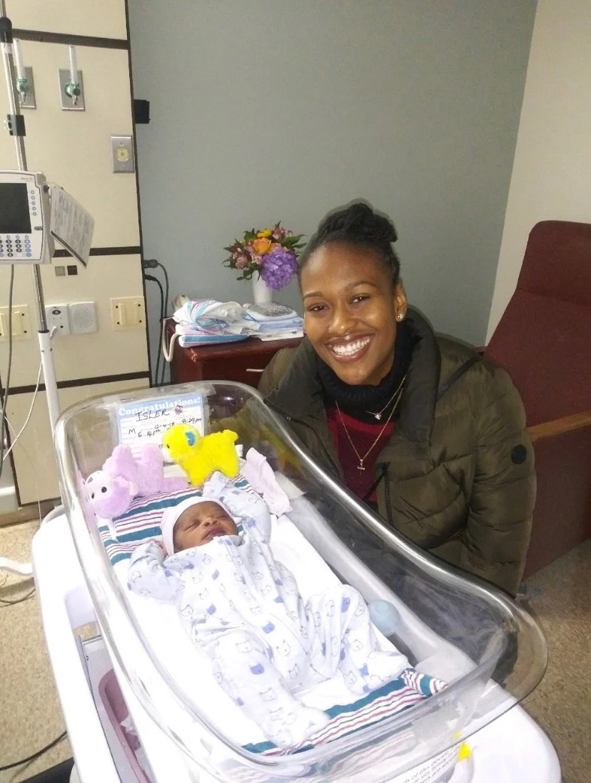 A smiling woman is standing next to a hospital bassinet containing a newborn baby. The baby is wearing a patterned onesie and is surrounded by small stuffed toys. Flowers are in the background.