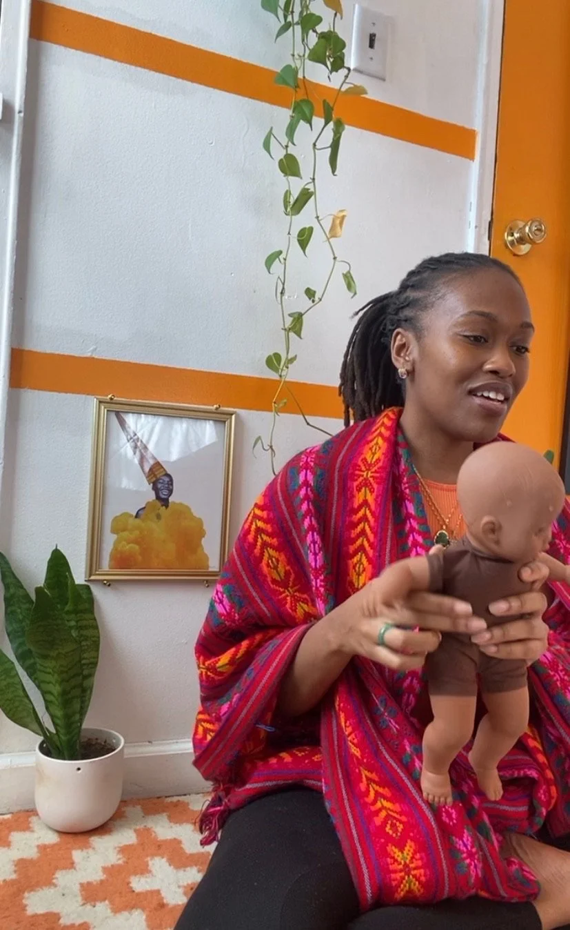 Person in colorful shawl holding a baby doll, with a framed picture and a houseplant in the background.