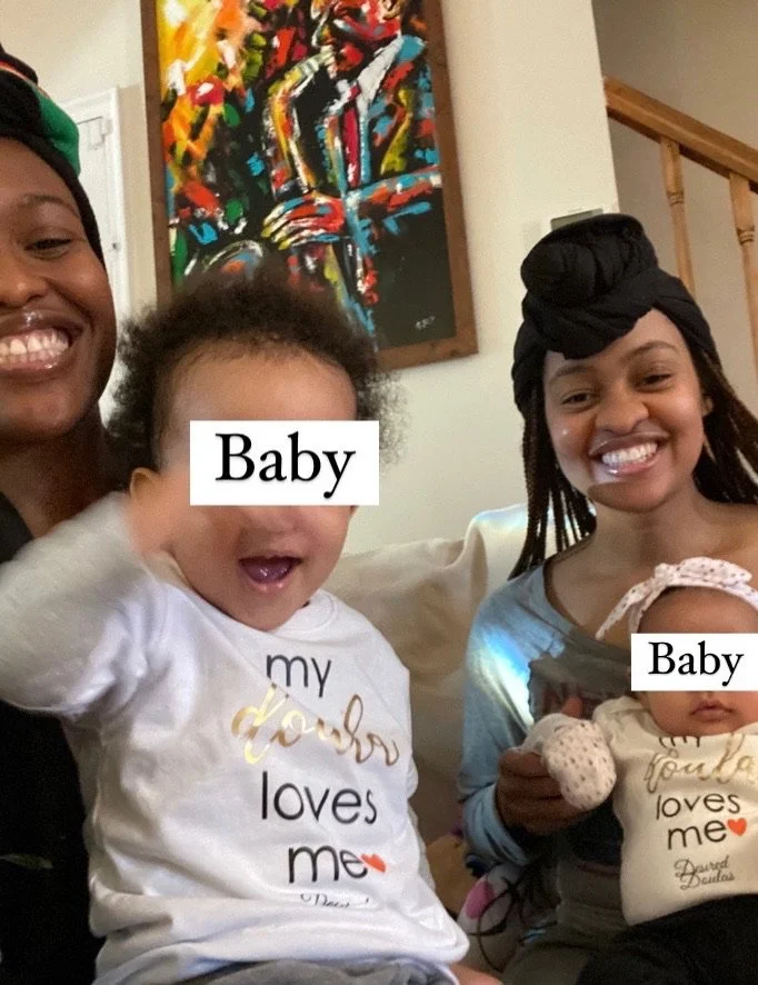 Two smiling women holding babies in a living room with colorful artwork on the wall.