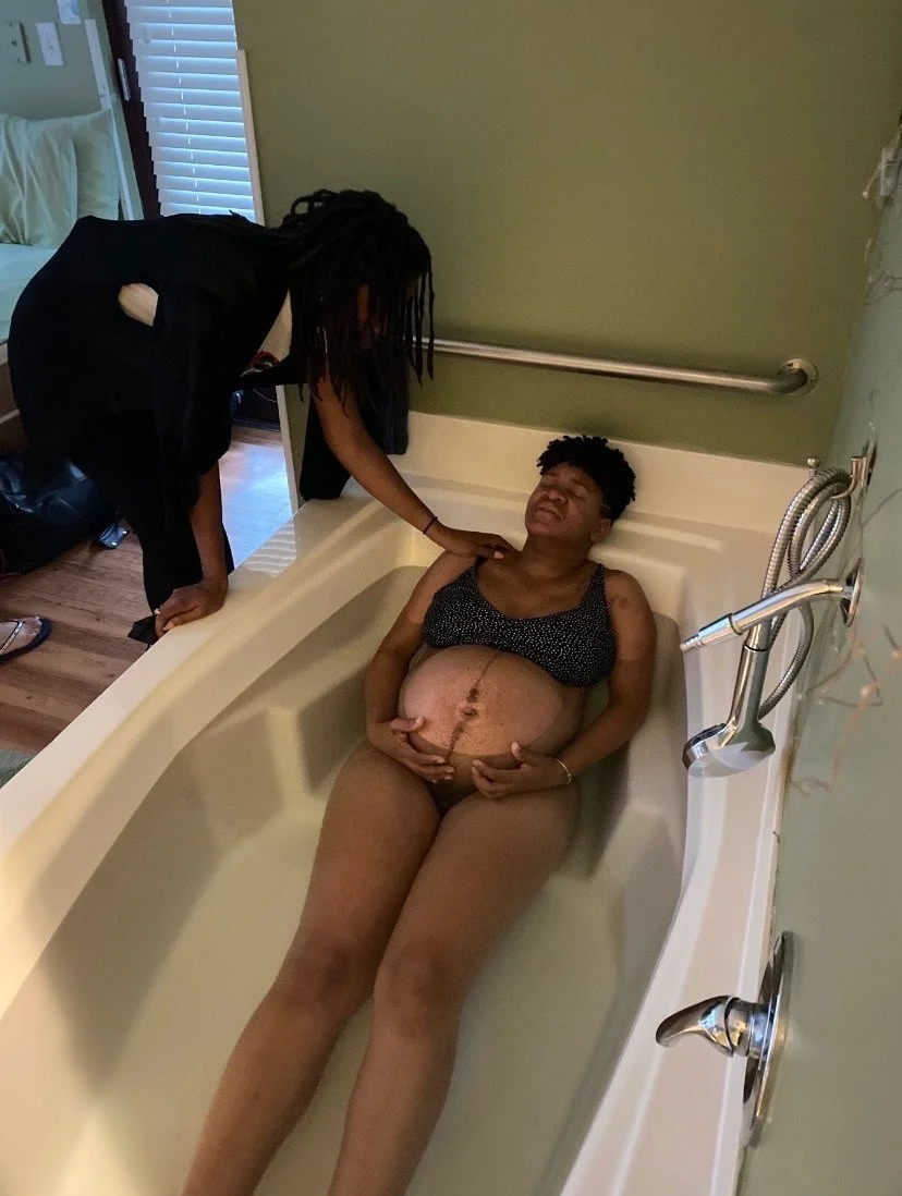 Pregnant person sitting in bathtub with support from another person, holding belly, appearing in labor.