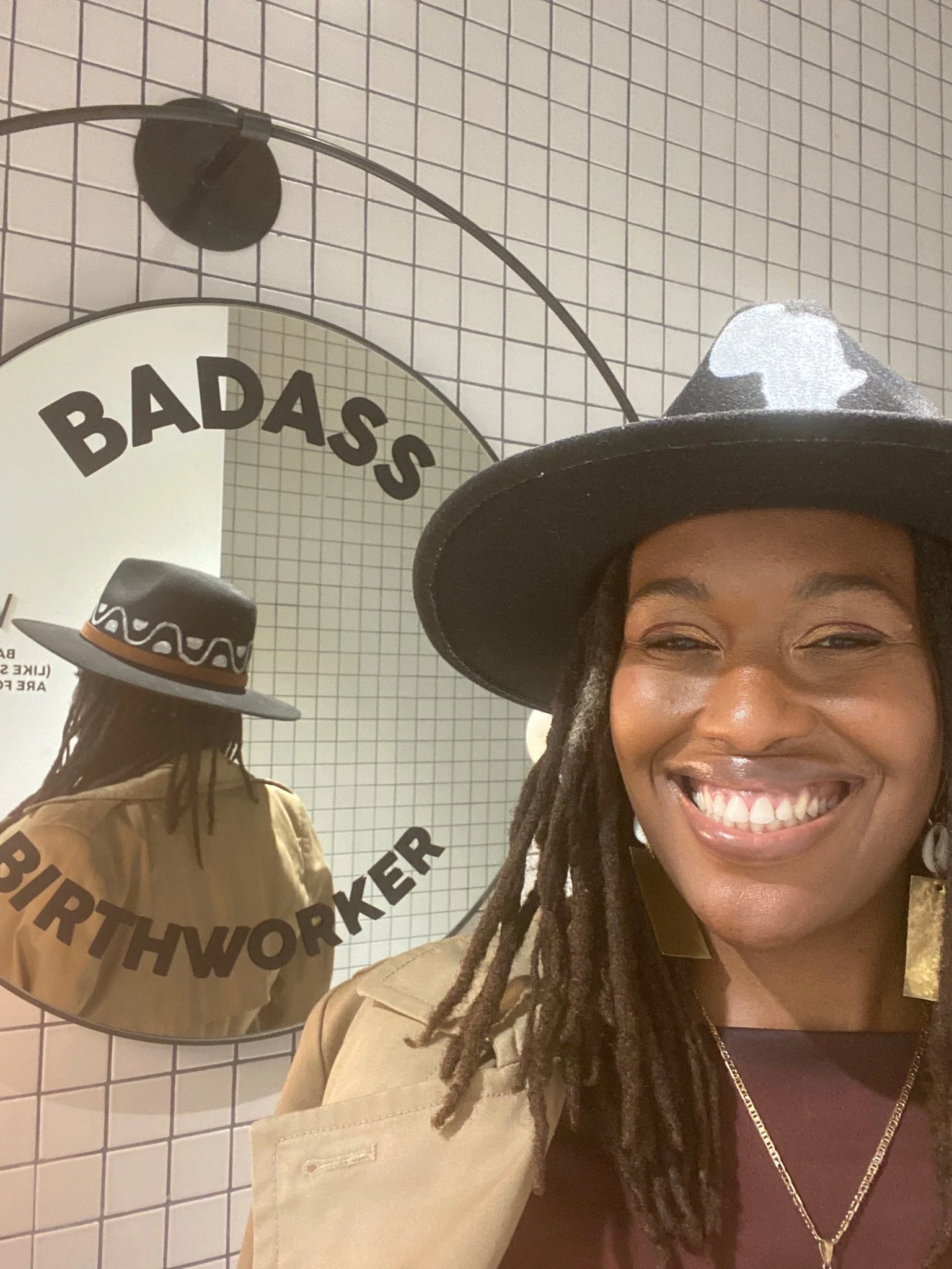 Person smiling in front of a circular mirror with "BADASS BIRTHWORKER" written around it, wearing a wide-brimmed hat and a beige jacket, standing against a white tiled wall.