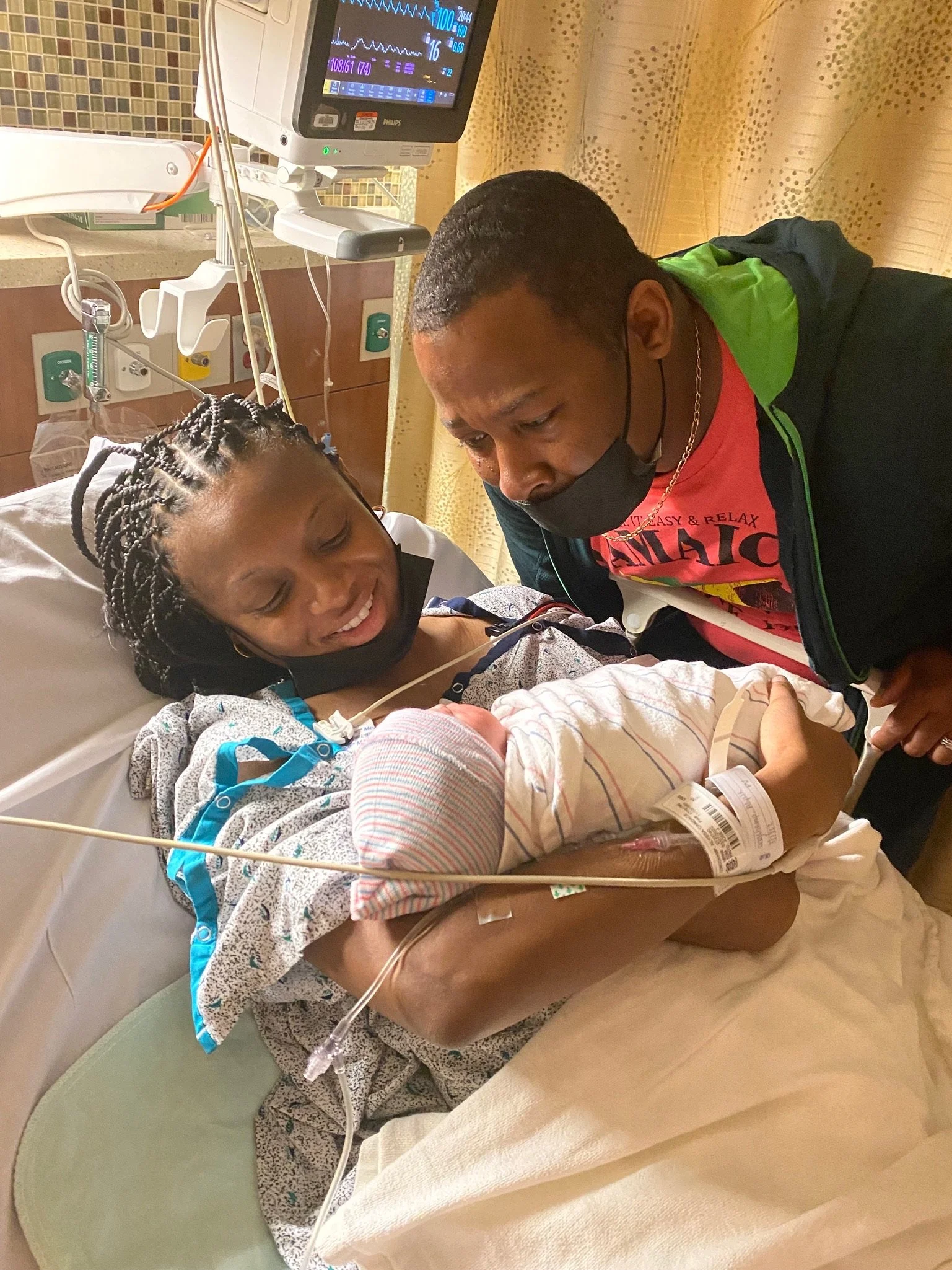 A mother holding a newborn baby in a hospital bed, with a man standing beside her. The mother is smiling, and both adults are wearing face masks under their chins. Medical equipment is visible in the background.