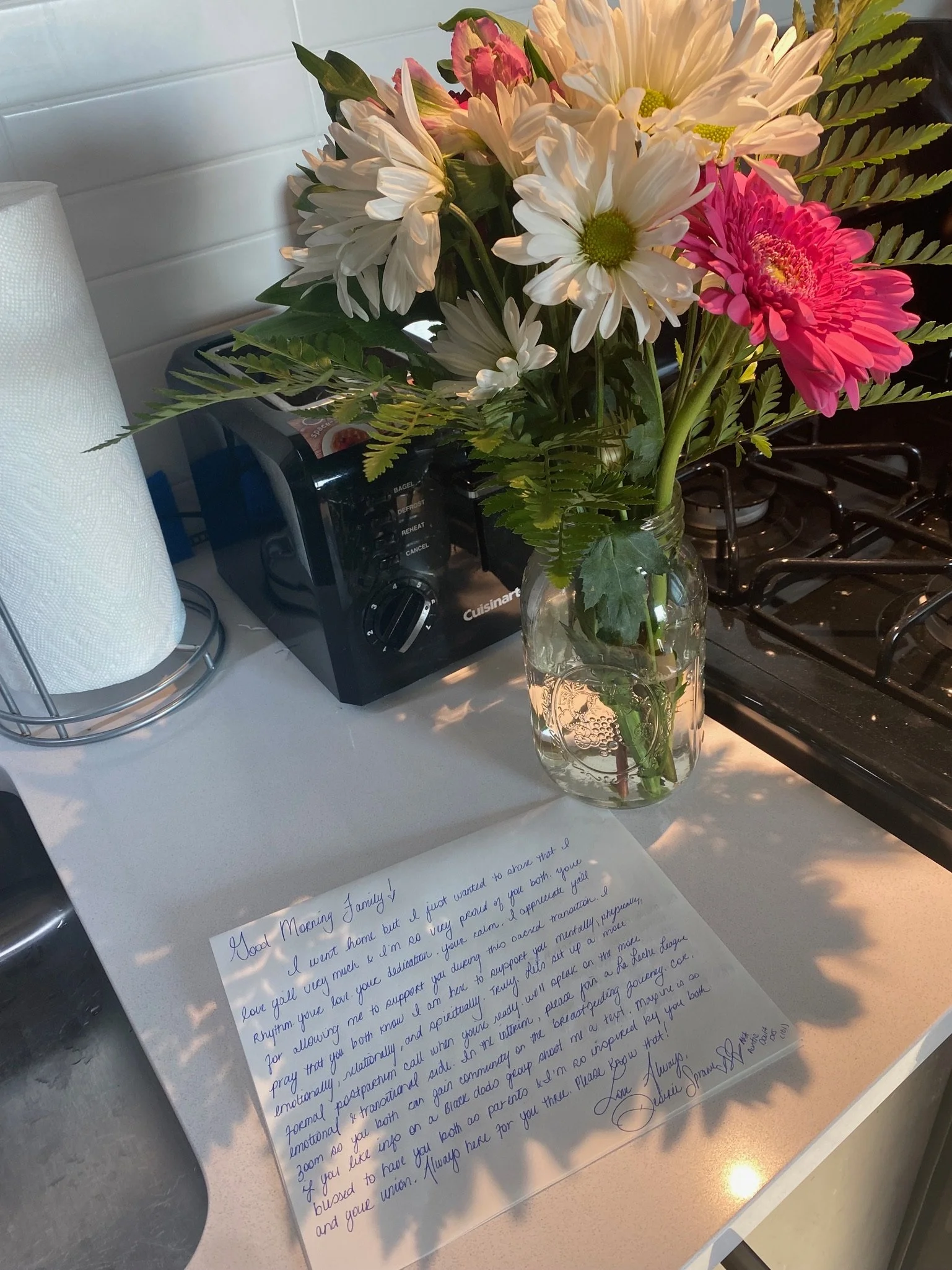 A bouquet of flowers in a mason jar on a kitchen counter next to a letter. The bouquet includes white and pink flowers with green leaves. A paper towel holder and toaster are on the counter beside the bouquet. The letter is handwritten in blue ink an