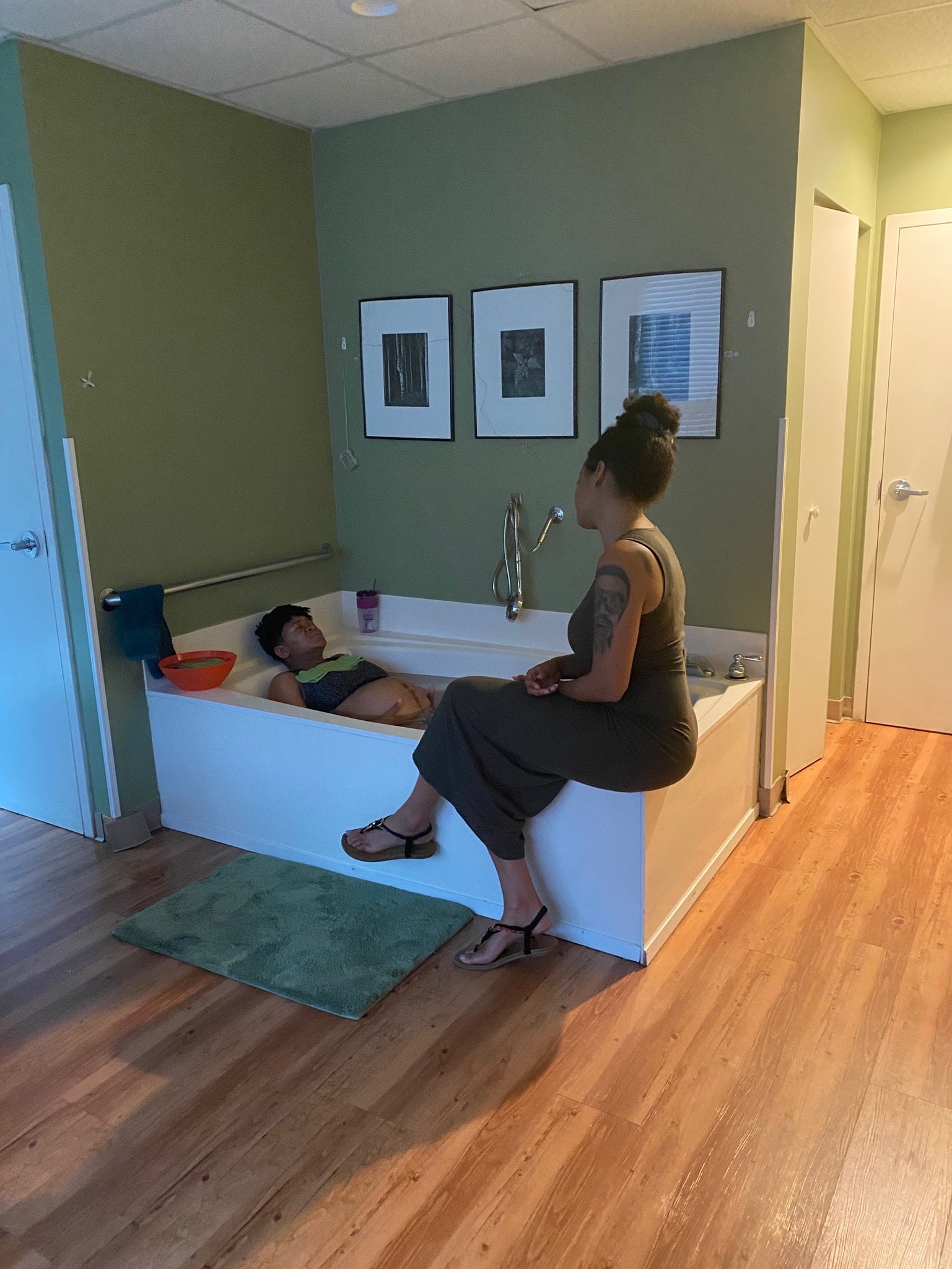 A person sitting on the edge of a bathtub talking to another person inside the tub in a dimly lit room with wooden floors and green walls, decorated with three framed pictures on the wall.