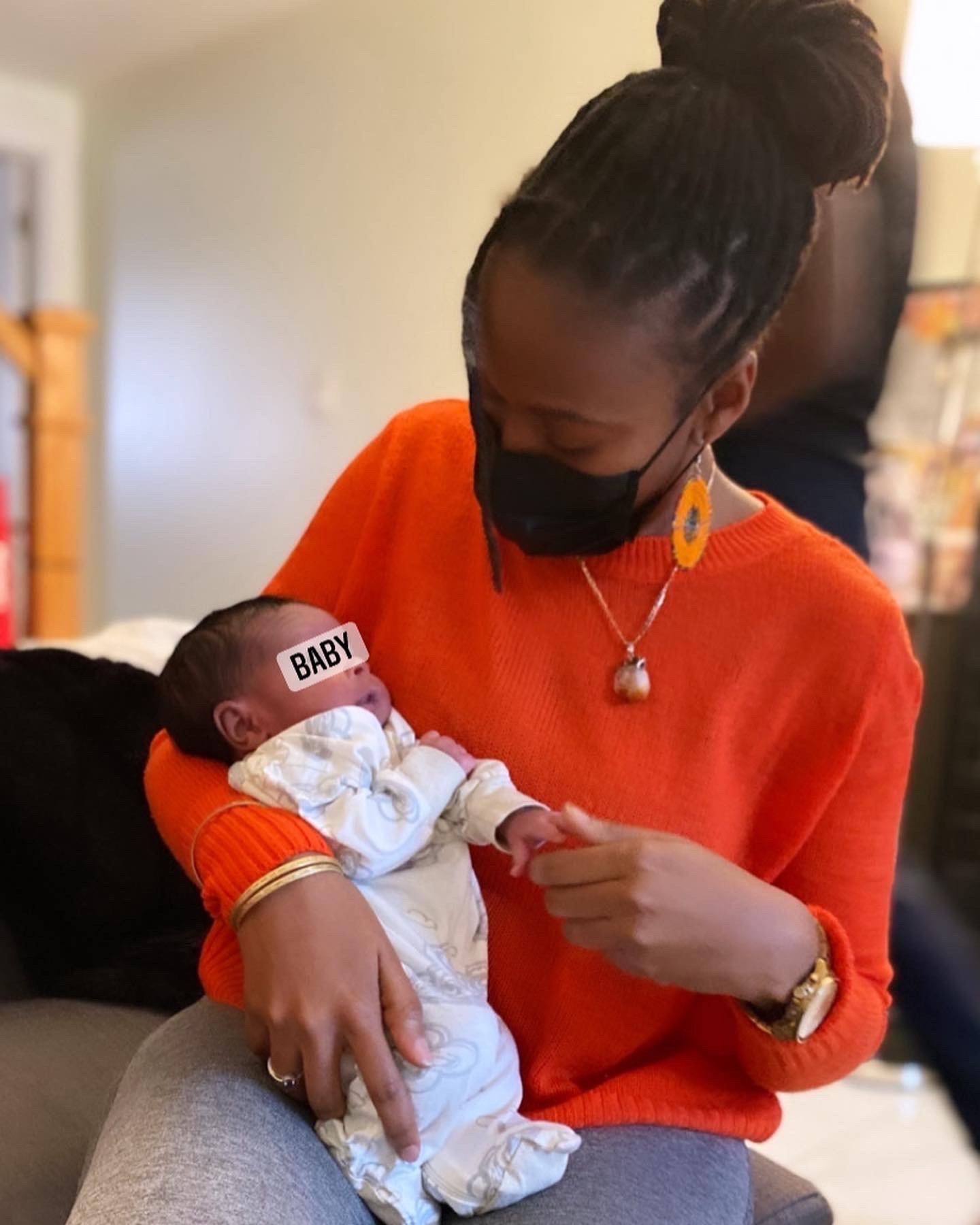 Woman in orange sweater holding a newborn baby, wearing a black mask and yellow earrings.