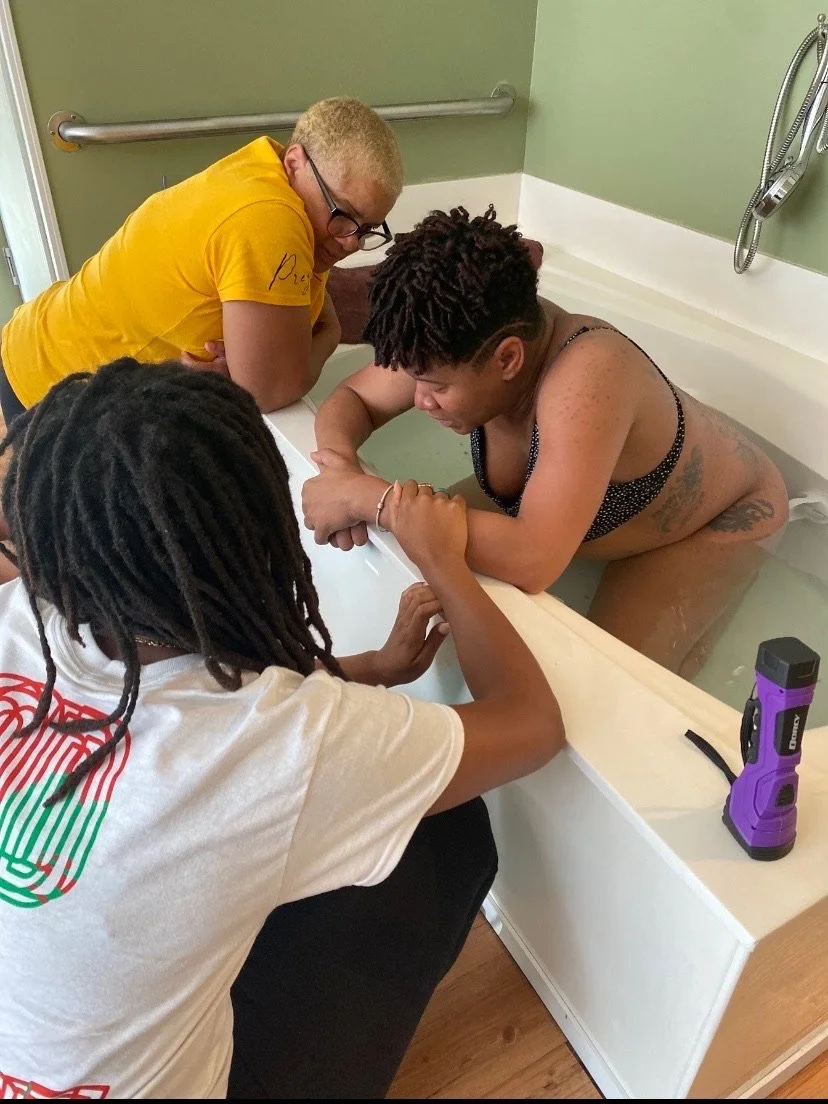 A person in a black swimsuit is leaning on the edge of a bathtub during a water birth, attended by two individuals, one wearing a yellow shirt and the other with long hair.