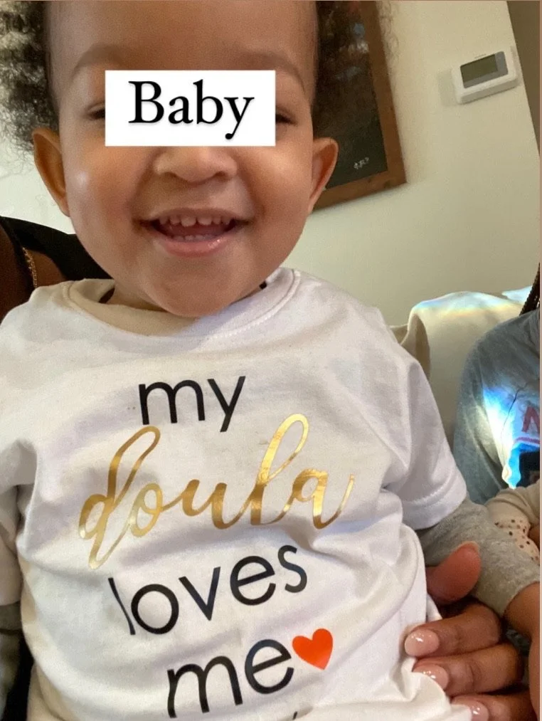 Smiling baby wearing a white shirt with the text 'my doula loves me' and a heart symbol, sitting on someone's lap.
