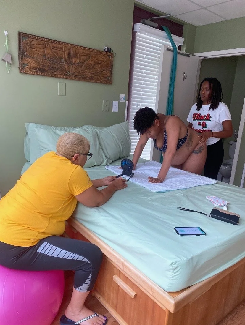 A pregnant woman kneels on a bed, supported by another woman. A third woman sits on an exercise ball, holding a device. The room has light green walls, a wooden carving, and white blinds.