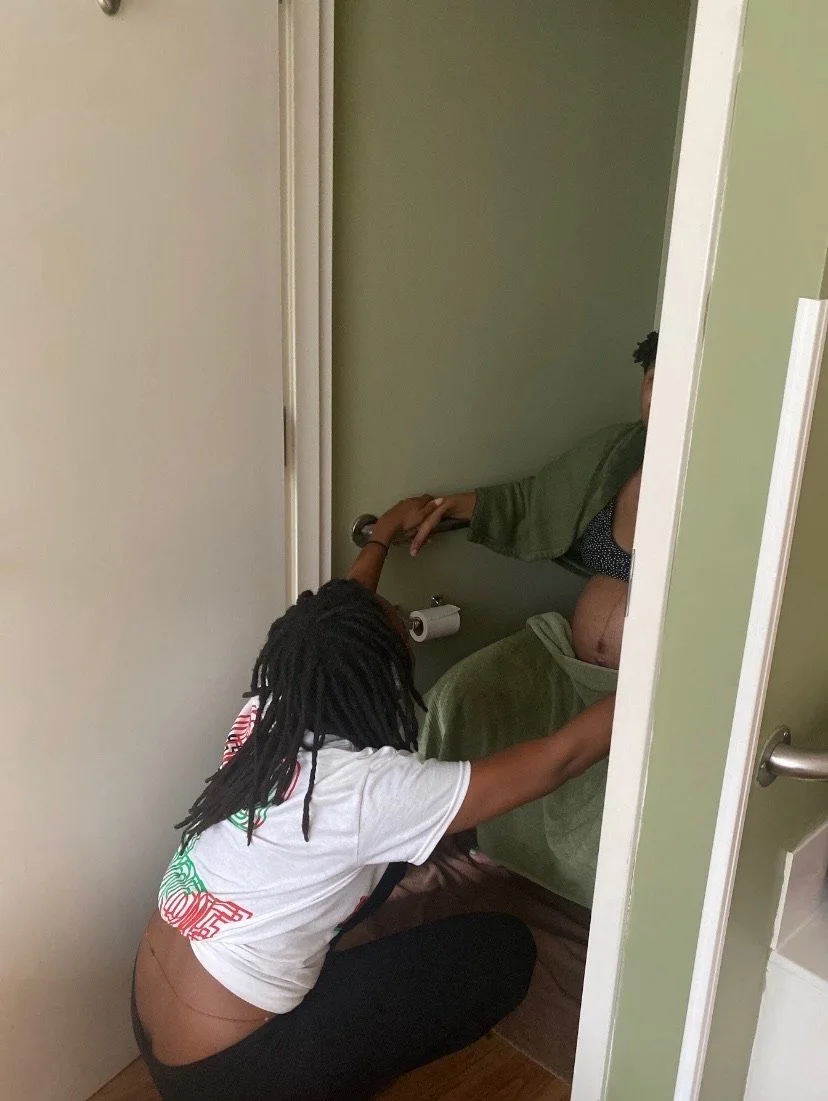 Two people are in a bathroom. One person is sitting, possibly on a toilet, covered with a green robe. The other person is kneeling, holding the first person's hand, dressed in black pants and a white t-shirt with red and green text. A roll of toilet 