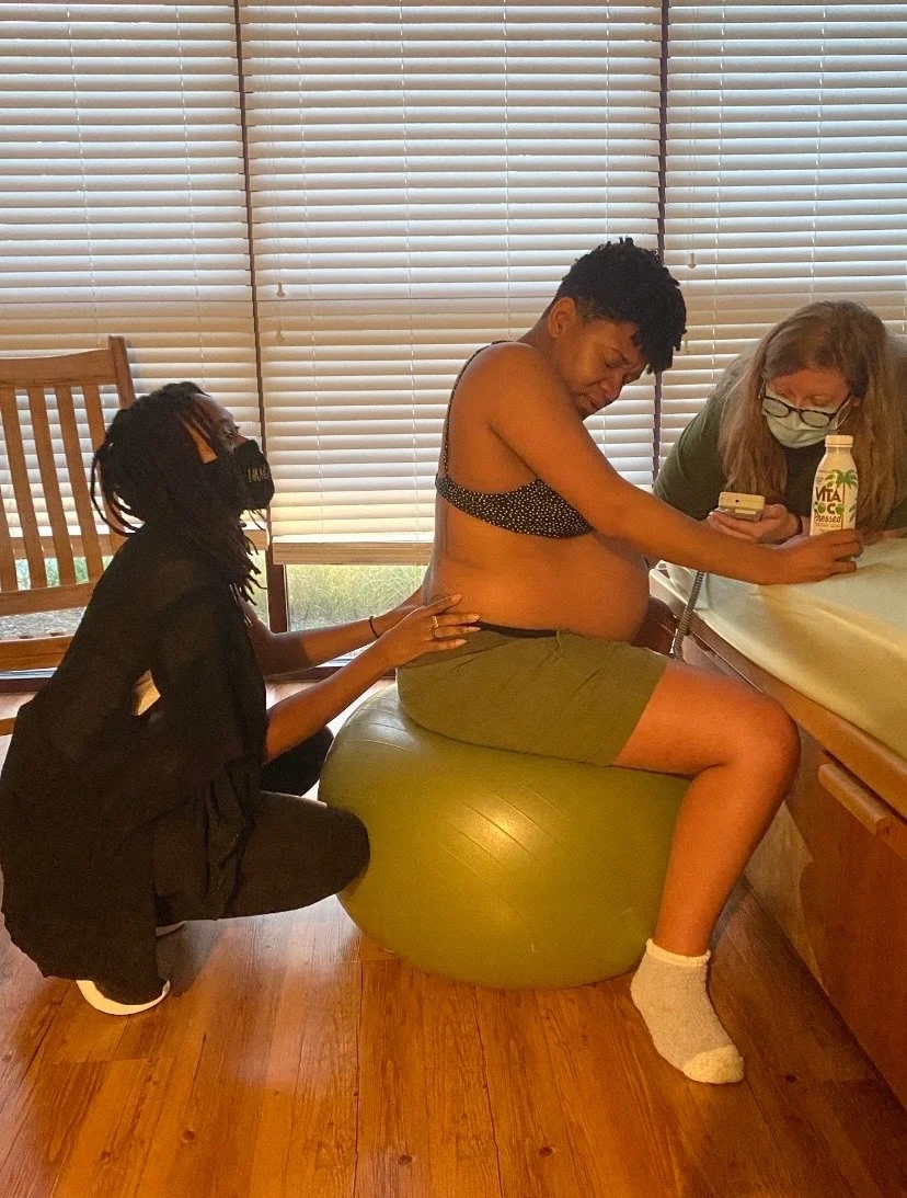 A pregnant person sitting on an exercise ball, supported by two individuals in a room with wooden flooring and blinds.