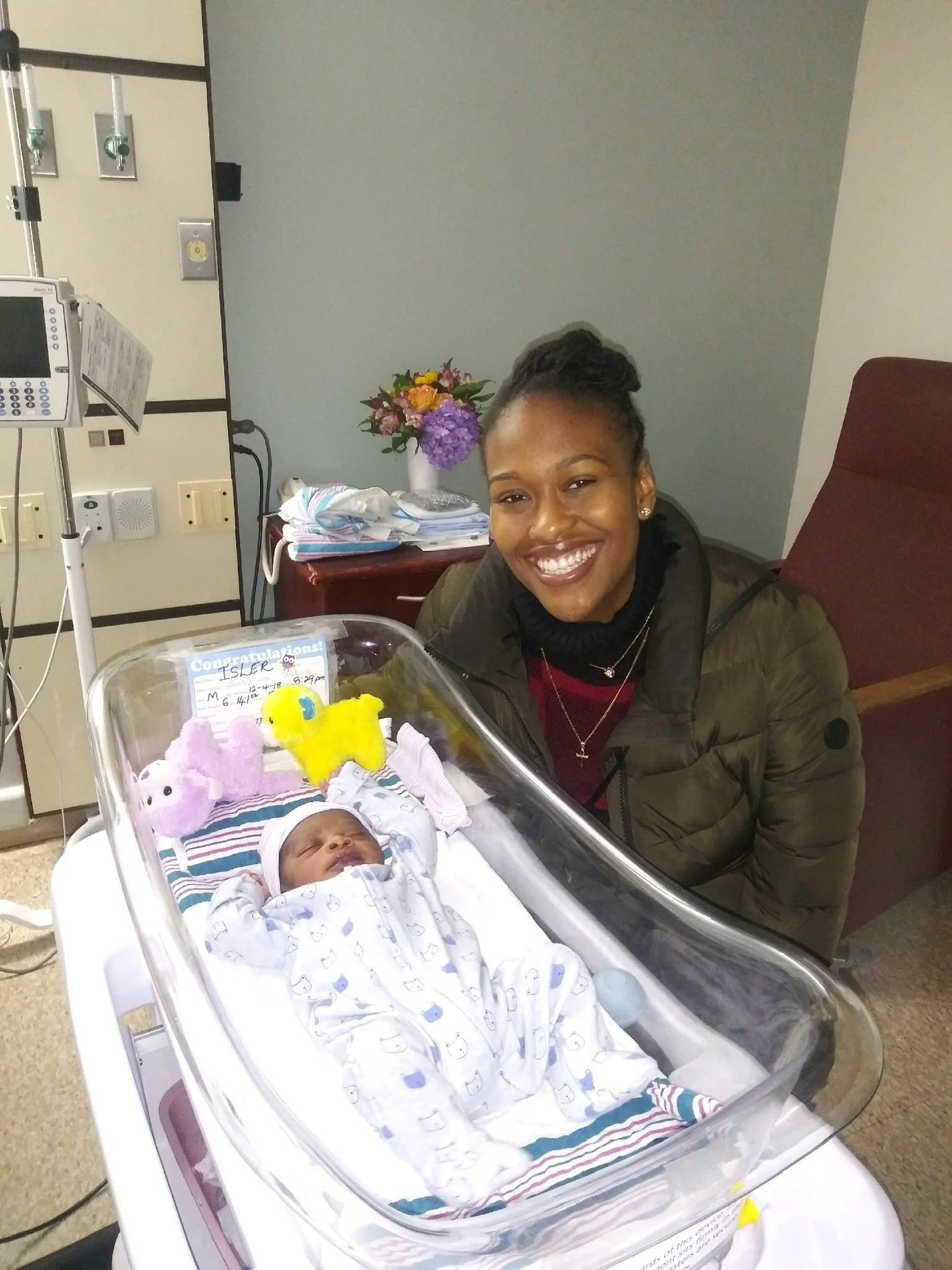 A woman smiling next to a newborn baby in a hospital bassinet. The infant is wearing a light-colored onesie with patterns and a cap, surrounded by plush toys. A bouquet of flowers and medical equipment are visible in the background.