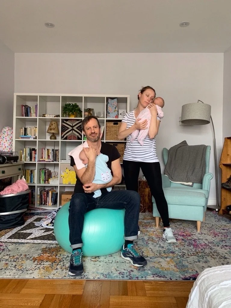 A man and a woman holding two babies in a cozy living room. The man is sitting on an exercise ball, holding a baby in blue, while the woman is standing and holding a baby in pink. Behind them is a bookshelf filled with books and decor, and an armchai