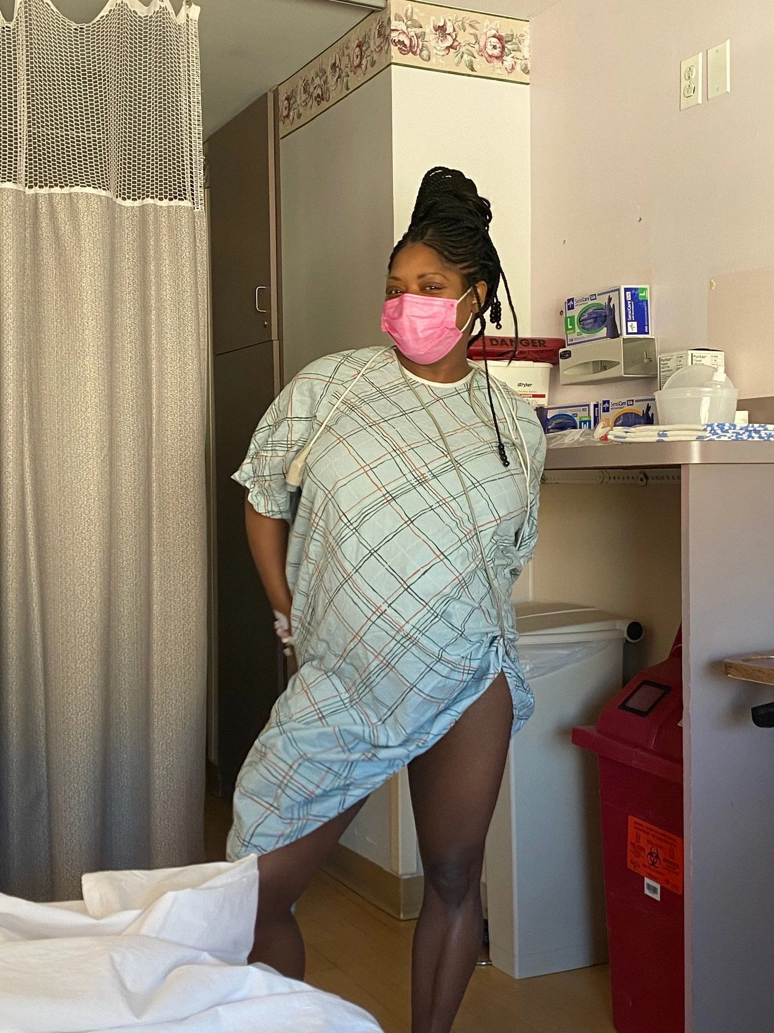 Person wearing a hospital gown and pink face mask standing in a medical room with medical supplies and a privacy curtain in the background.