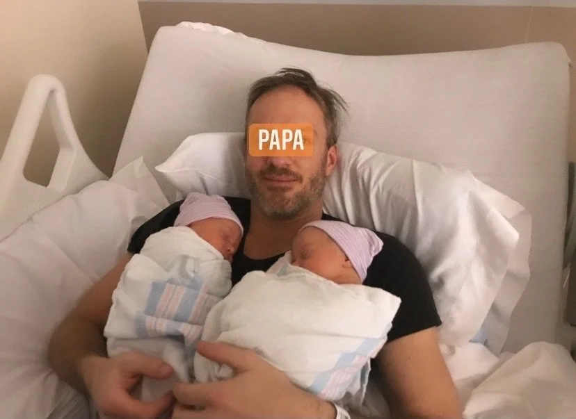 Man lying in hospital bed holding twin newborn babies wrapped in blankets.