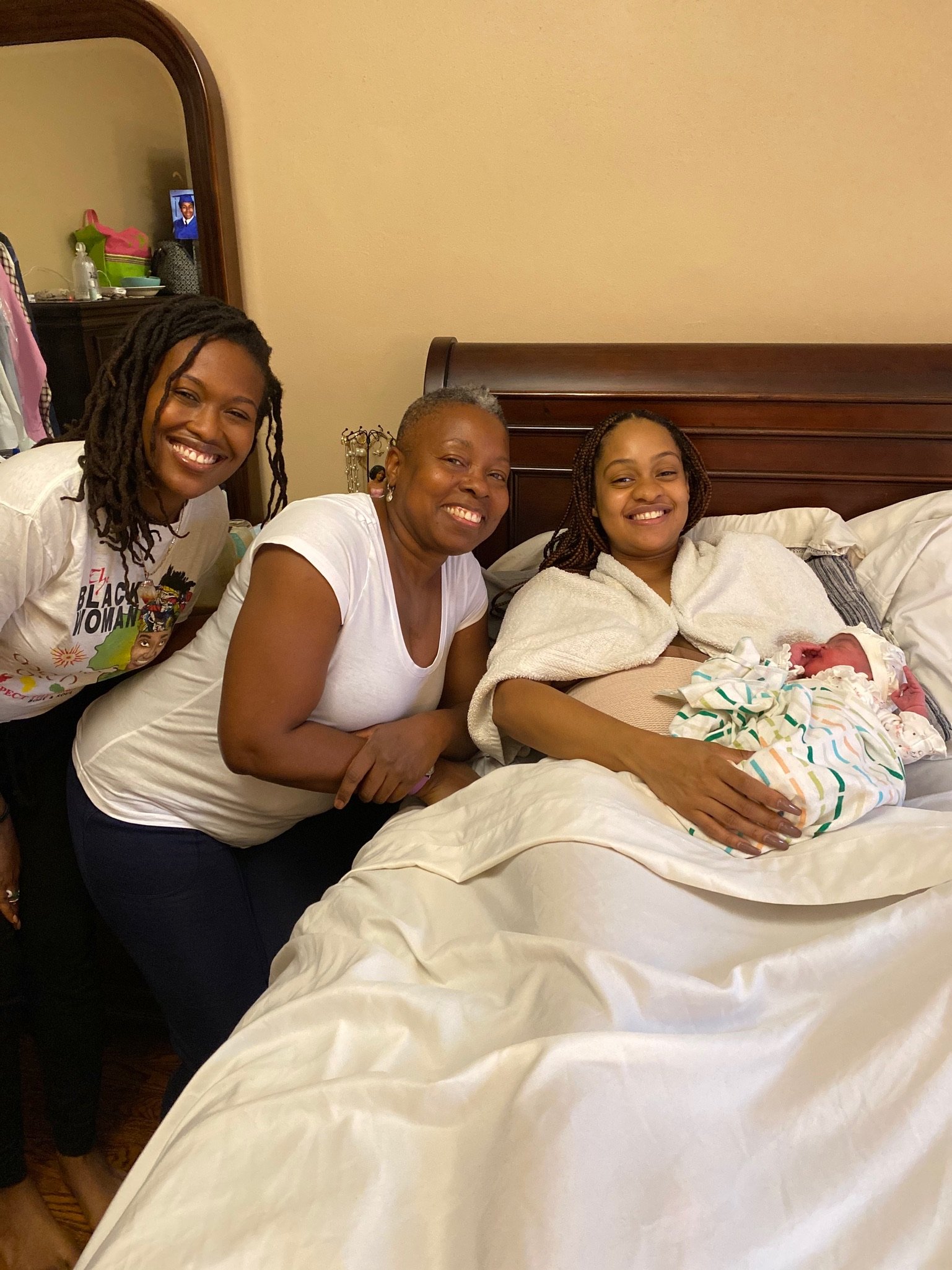 Three women smiling next to a new mother holding a baby wrapped in a striped blanket, lying on a bed in a cozy room.