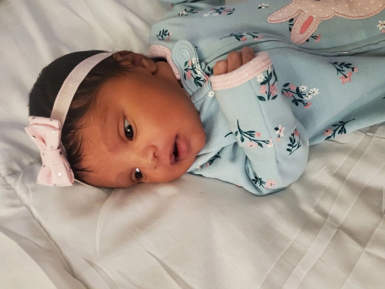 Newborn baby wearing a floral onesie and a pink bow headband, lying on a white blanket.