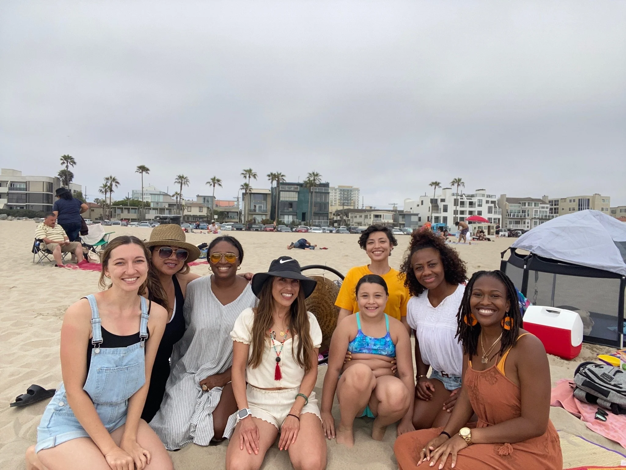 Group of women and a child smiling on a beach, seated on sand with beach houses and palm trees in the background.