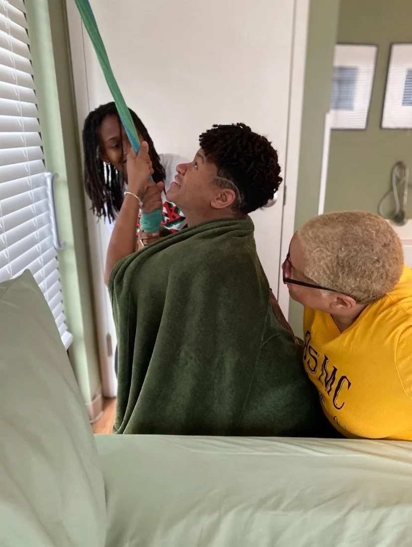 Person pulling on green rope with assistance, draped in a green blanket, in a room with a bed and closed blinds.