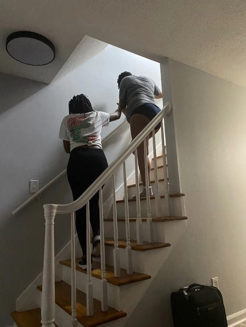 Two people ascending a staircase in a dimly lit interior, with a suitcase at the base of the stairs.