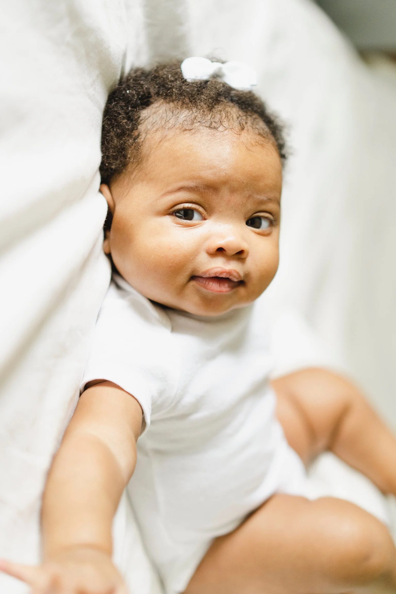 A baby in a white onesie with a small white bow on its head, sitting on a bed and looking at the camera.