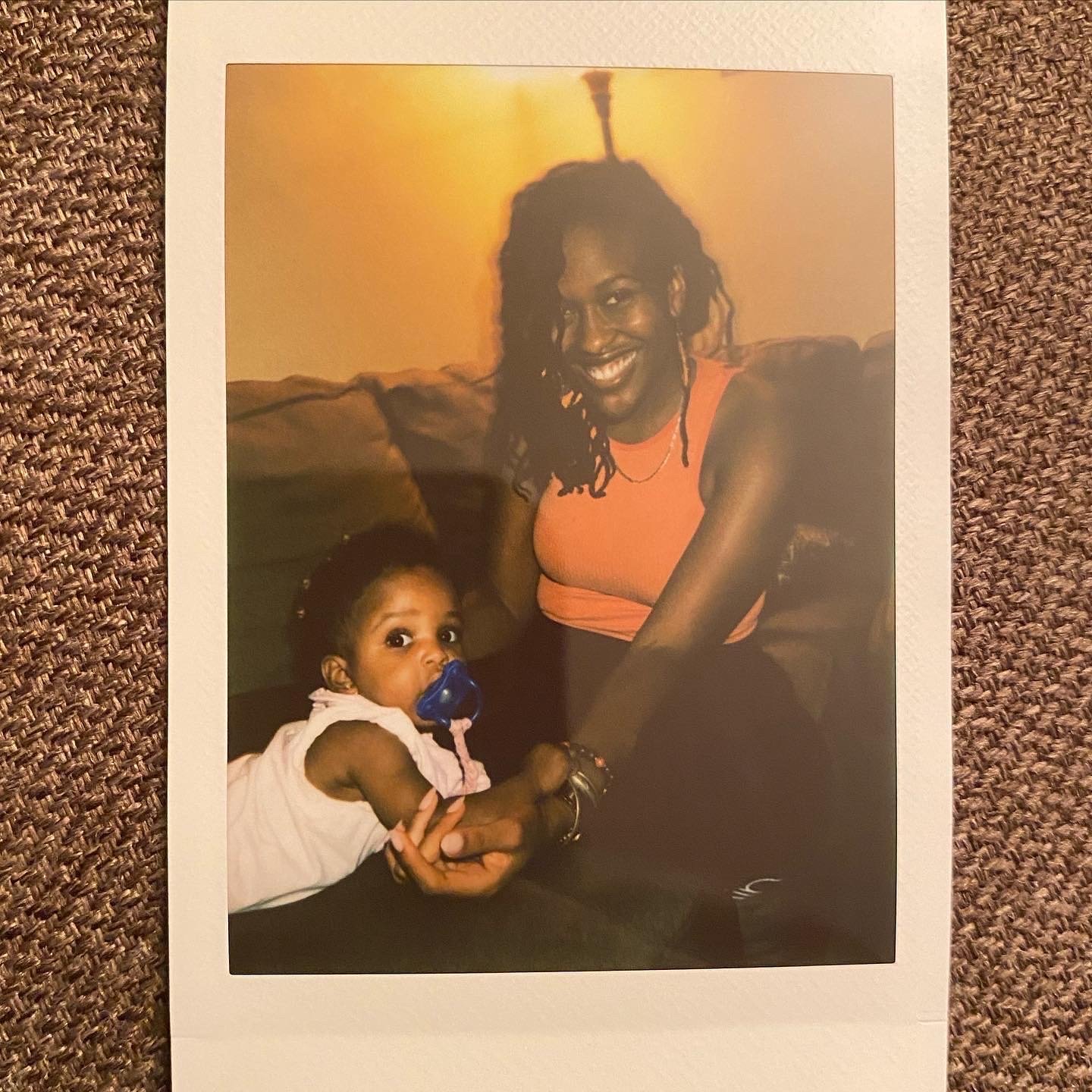 A smiling woman in an orange tank top sits on a couch, holding hands with a baby who has a blue pacifier. The background is softly lit, creating a warm atmosphere.