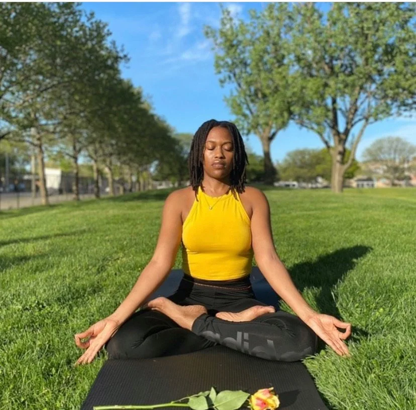 A woman practicing yoga in a lotus position on a mat outdoors, wearing a yellow top and black pants, with a single flower on the grass nearby.