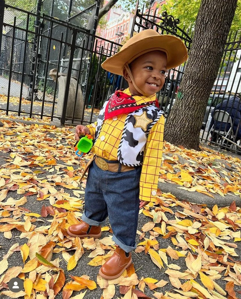 A child dressed in a cowboy costume with a sheriff badge, yellow plaid shirt, and red bandana, standing on a sidewalk covered with autumn leaves.