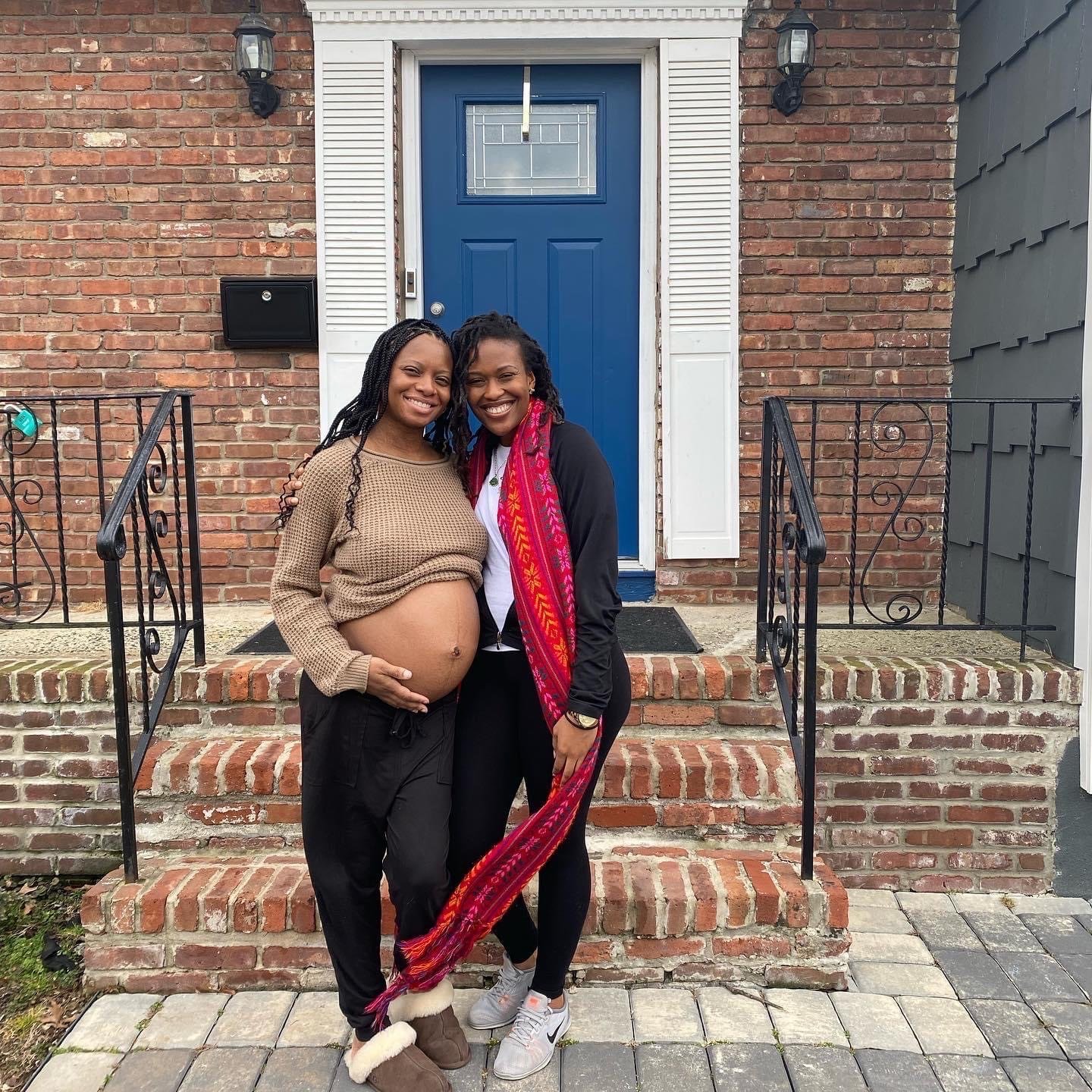 Two smiling women standing outside a house with a blue door. One woman is pregnant, showing her bare belly, and the other wears a vibrant scarf. They are on brick steps.