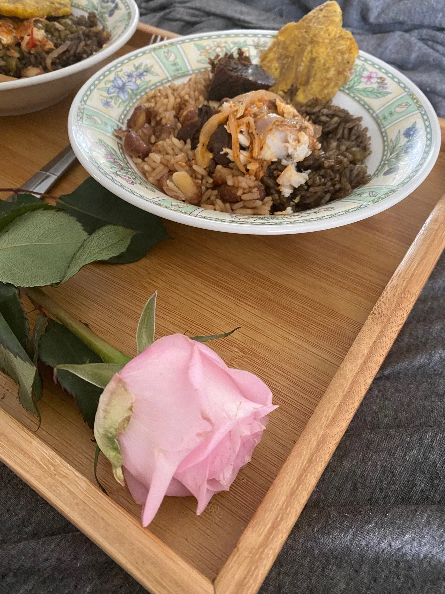 A bowl of rice with meats and a plantain chip on a wooden tray next to a pink rose.