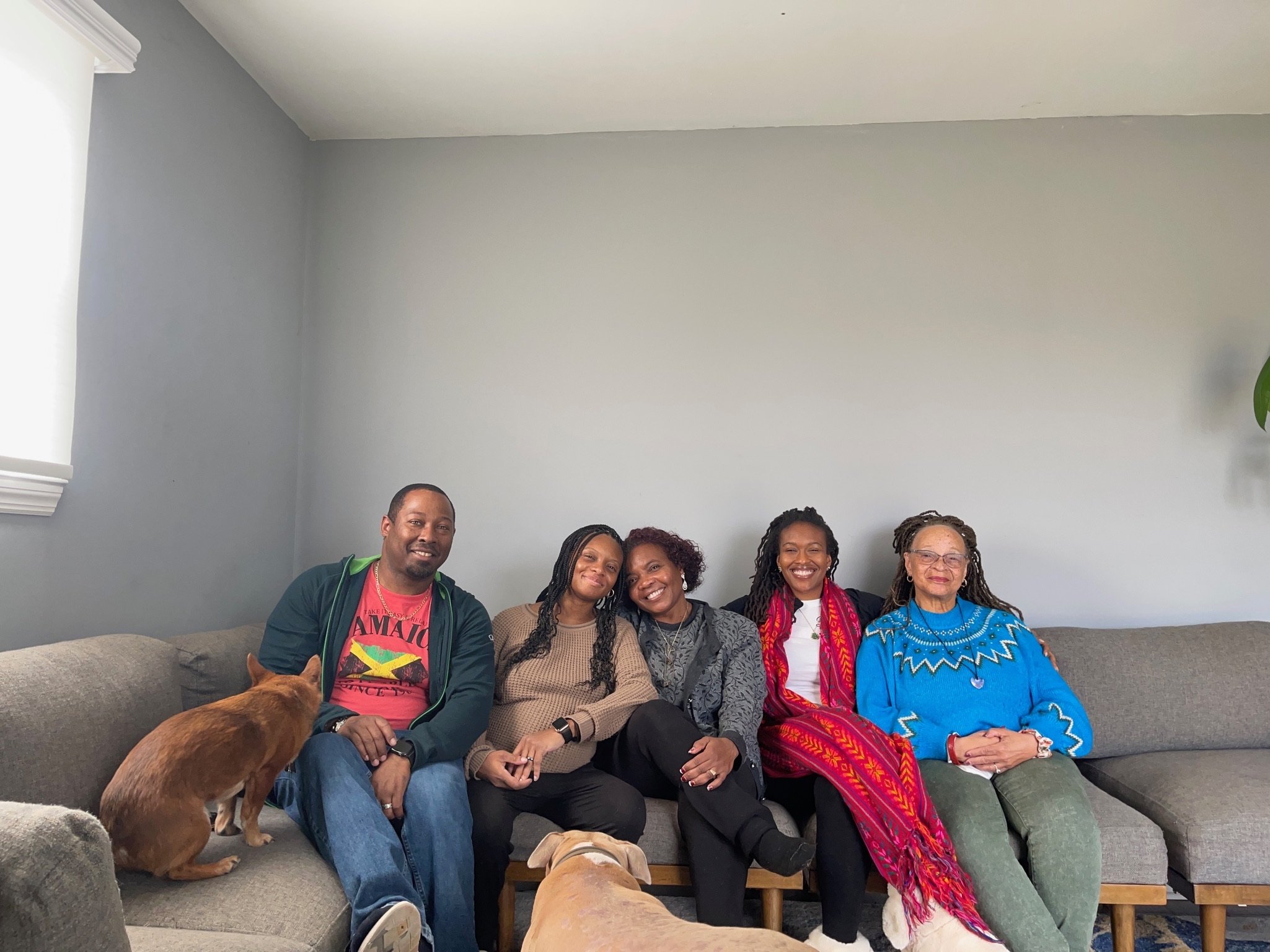A diverse group of five people sitting on a gray couch, smiling at the camera. Two dogs are also present, one on the couch and one sitting in front. The background features a gray wall and partially visible windows.