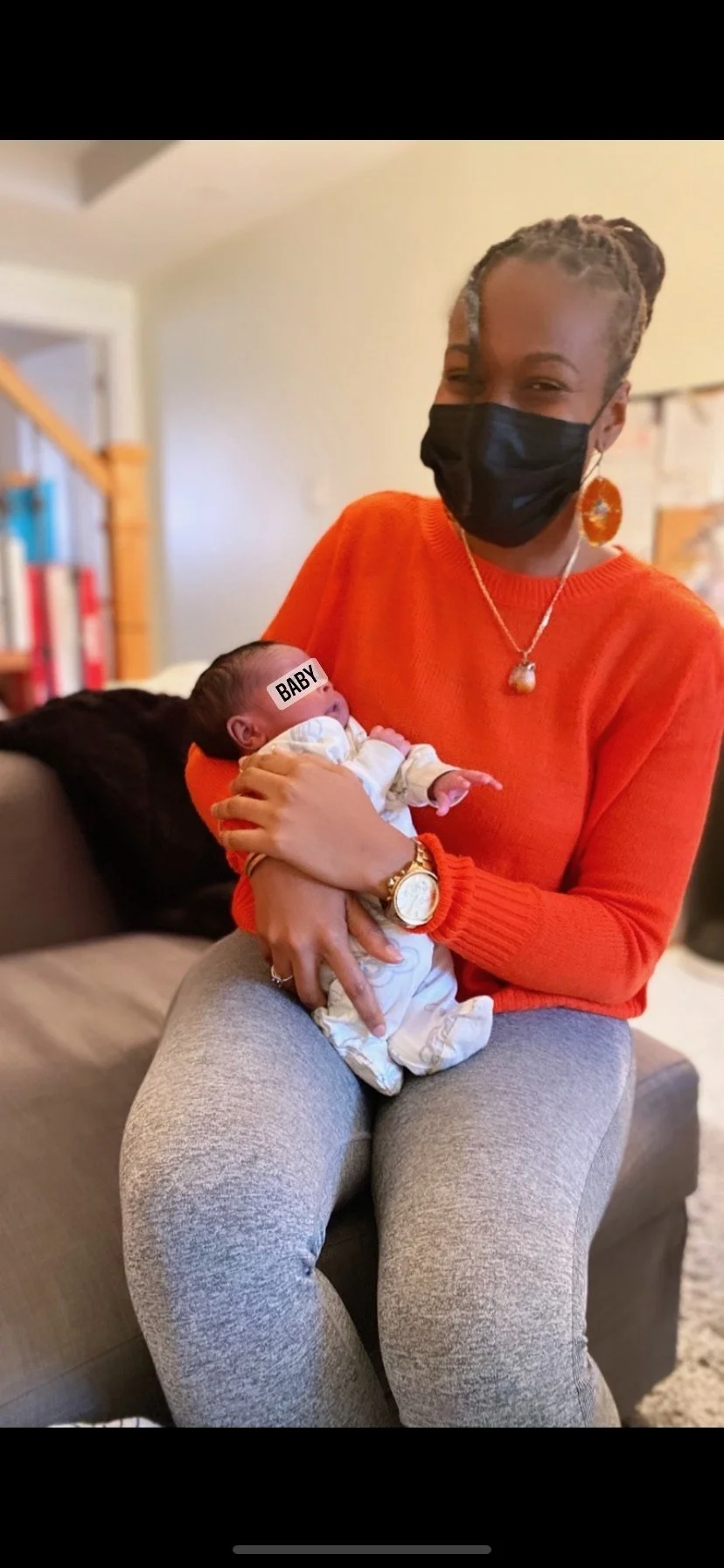 A person wearing an orange sweater and face mask is sitting on a couch holding a newborn baby dressed in white.