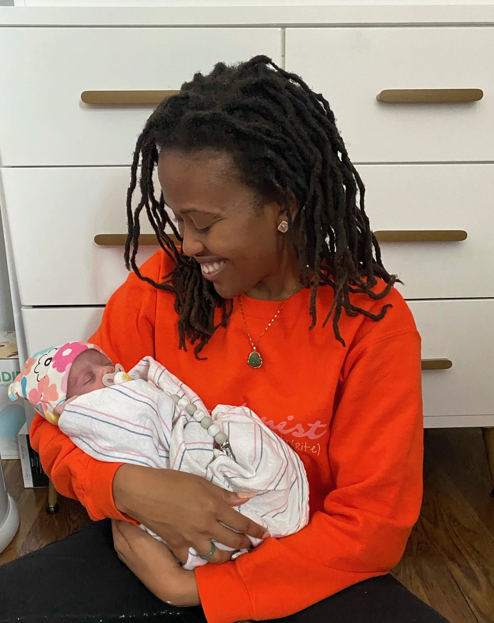 A person with long hair in an orange sweater smiling at a newborn baby wrapped in a blanket.