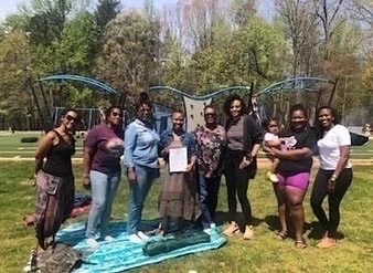A group of women standing on a lawn, posing for a picture in front of a blue playground. The women are dressed casually, and one is holding a baby. They are gathered on and around a blanket spread on the grass, enjoying a sunny day.