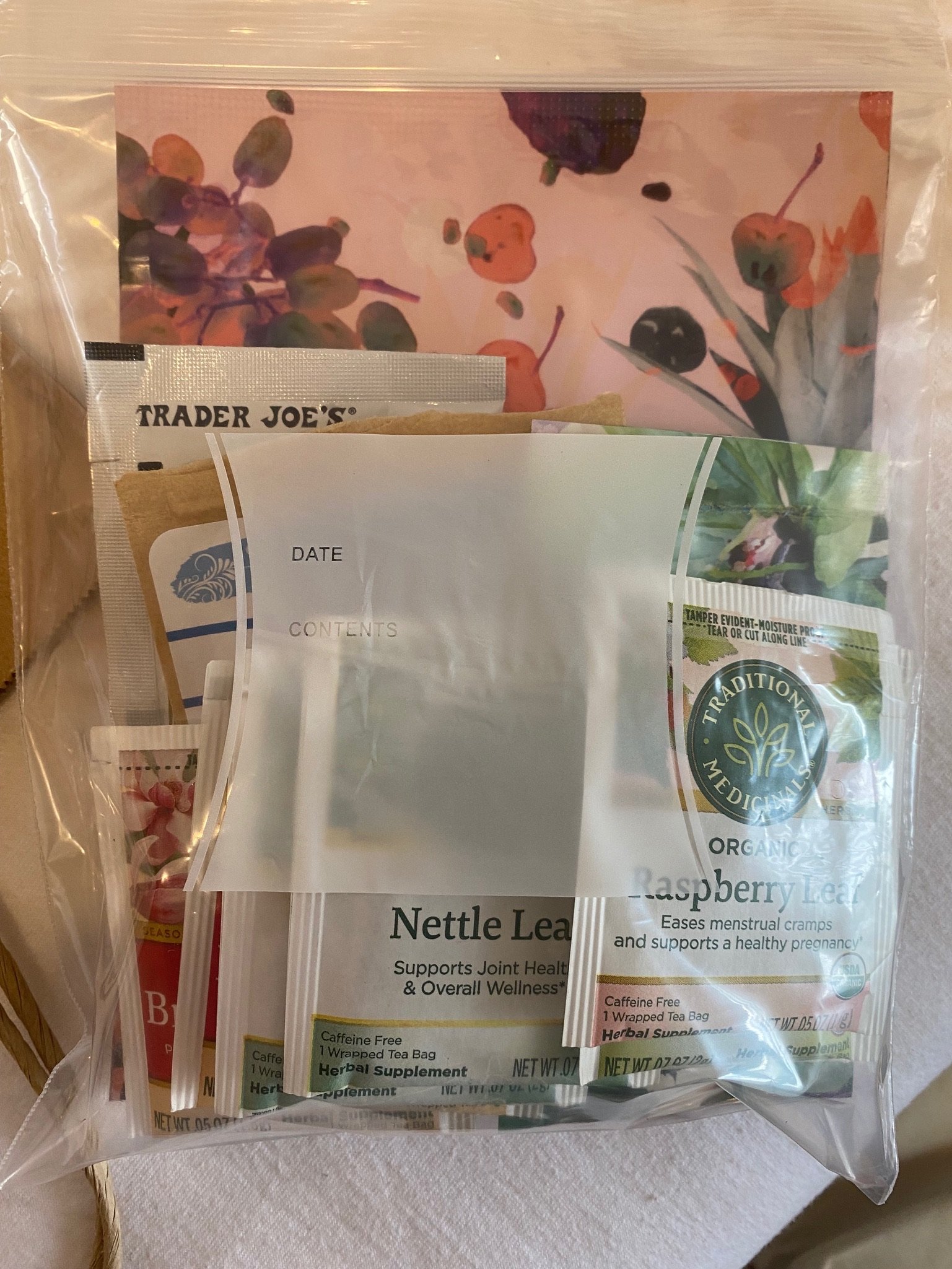 Plastic bag containing assorted tea packets, including Nettle Leaf and Raspberry Leaf teas, and a decorative card with a floral design.