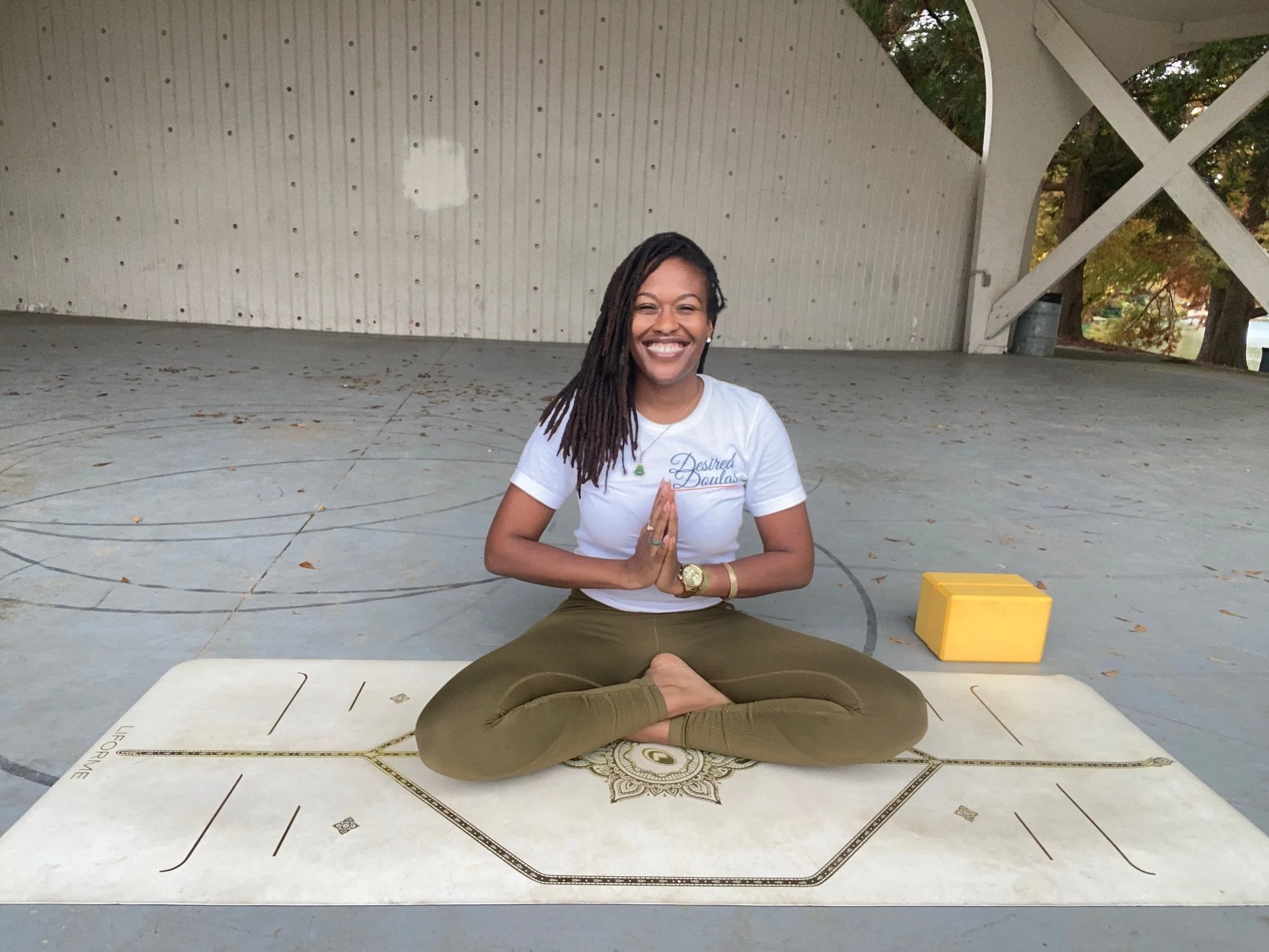 Person practicing yoga in outdoor pavilion, sitting in cross-legged position on a yoga mat, with hands in prayer pose and smiling. A yellow yoga block is placed nearby.