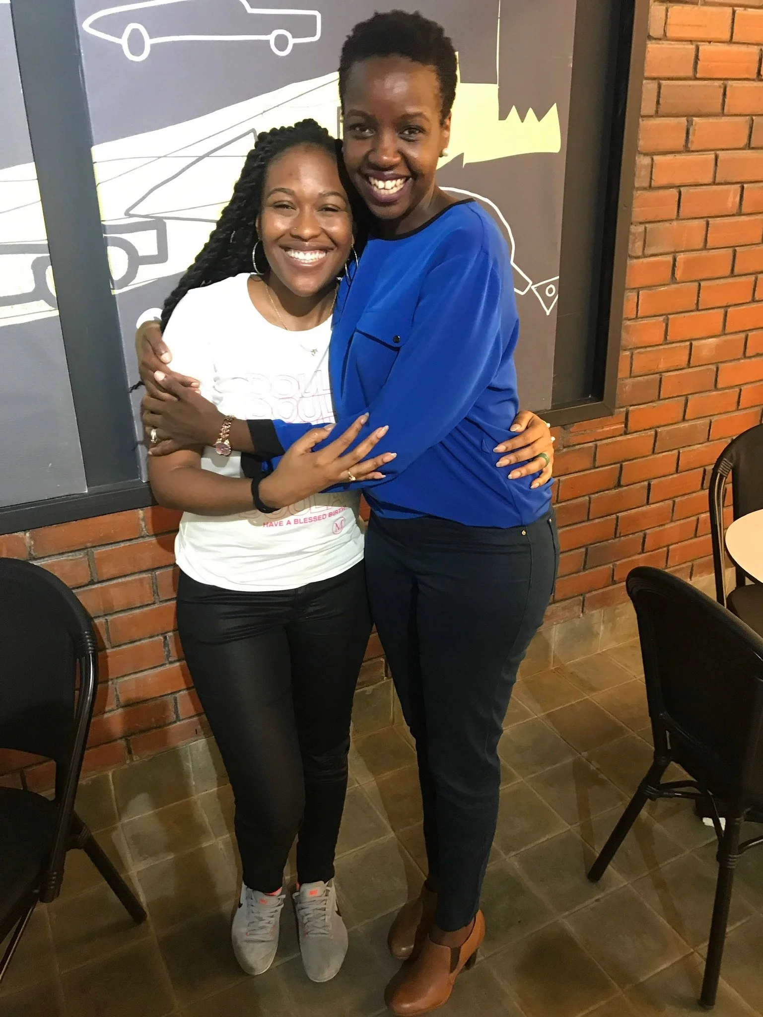 Two women smiling and hugging in an indoor setting with brick walls and chairs, one wearing a white t-shirt and the other in a blue top.