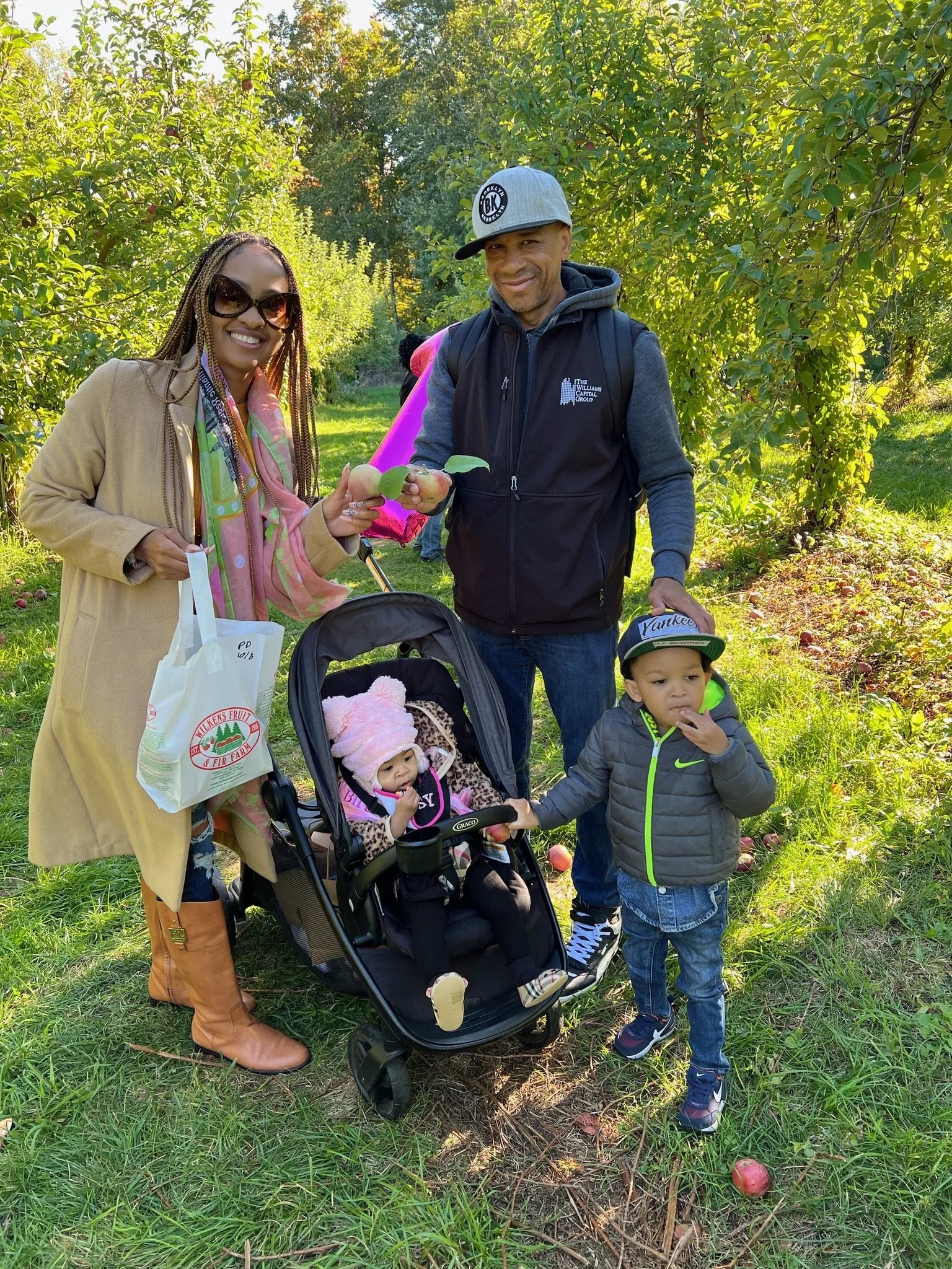 A family of four in an apple orchard; two adults smiling and holding apples, a baby in a stroller wearing a pink hat, and a young child eating an apple.
