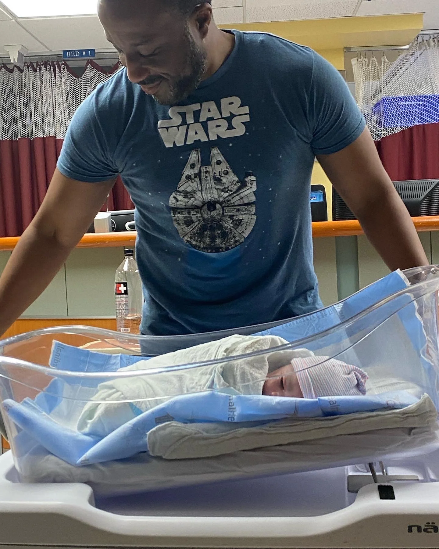 Man wearing Star Wars t-shirt looking at newborn baby in hospital bassinet.