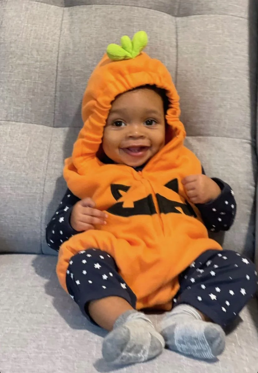 Smiling baby in a pumpkin costume with star-patterned pajamas on a gray chair.