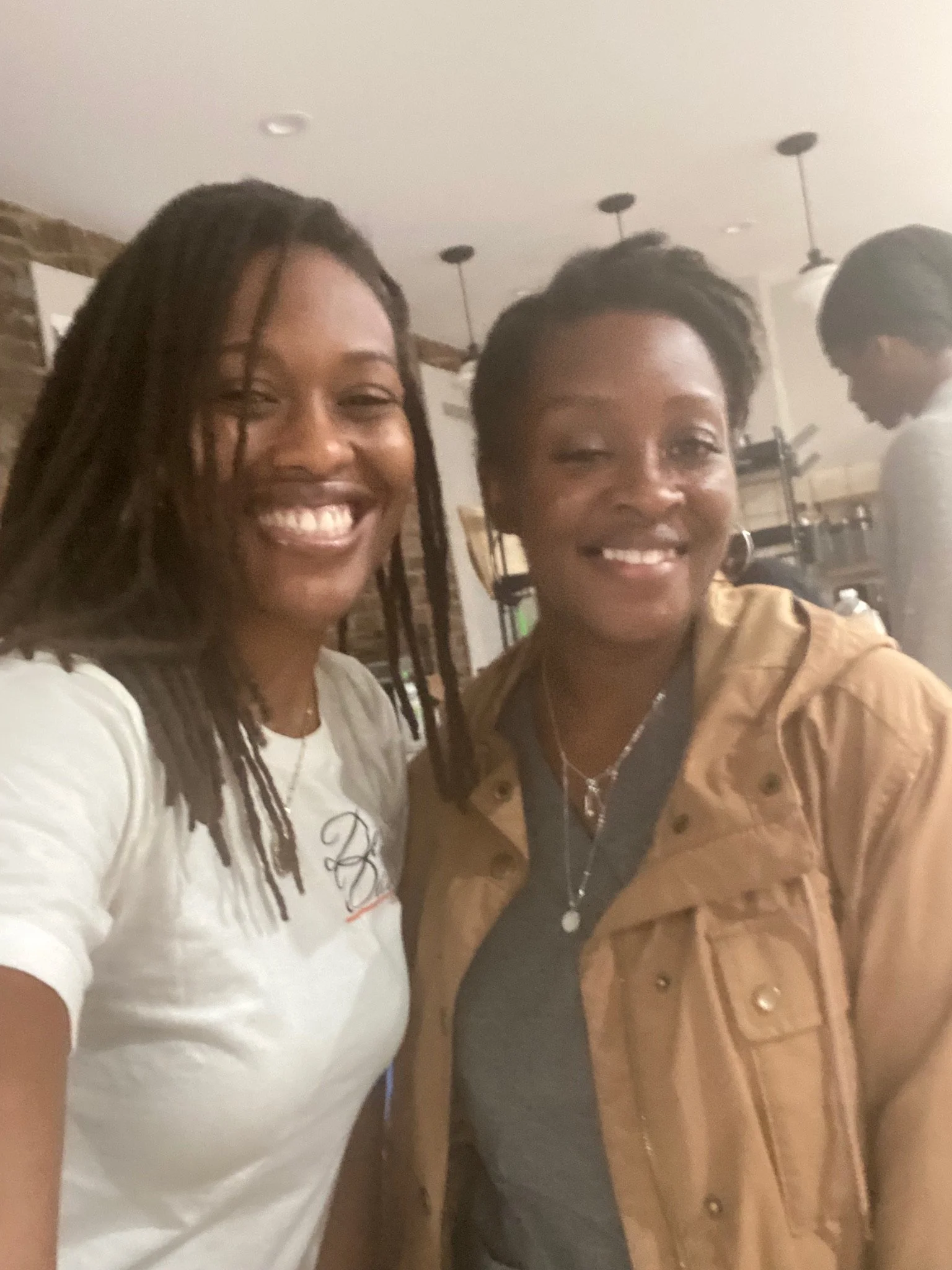 Two people smiling indoors, one wearing a white shirt, and the other wearing a brown jacket.