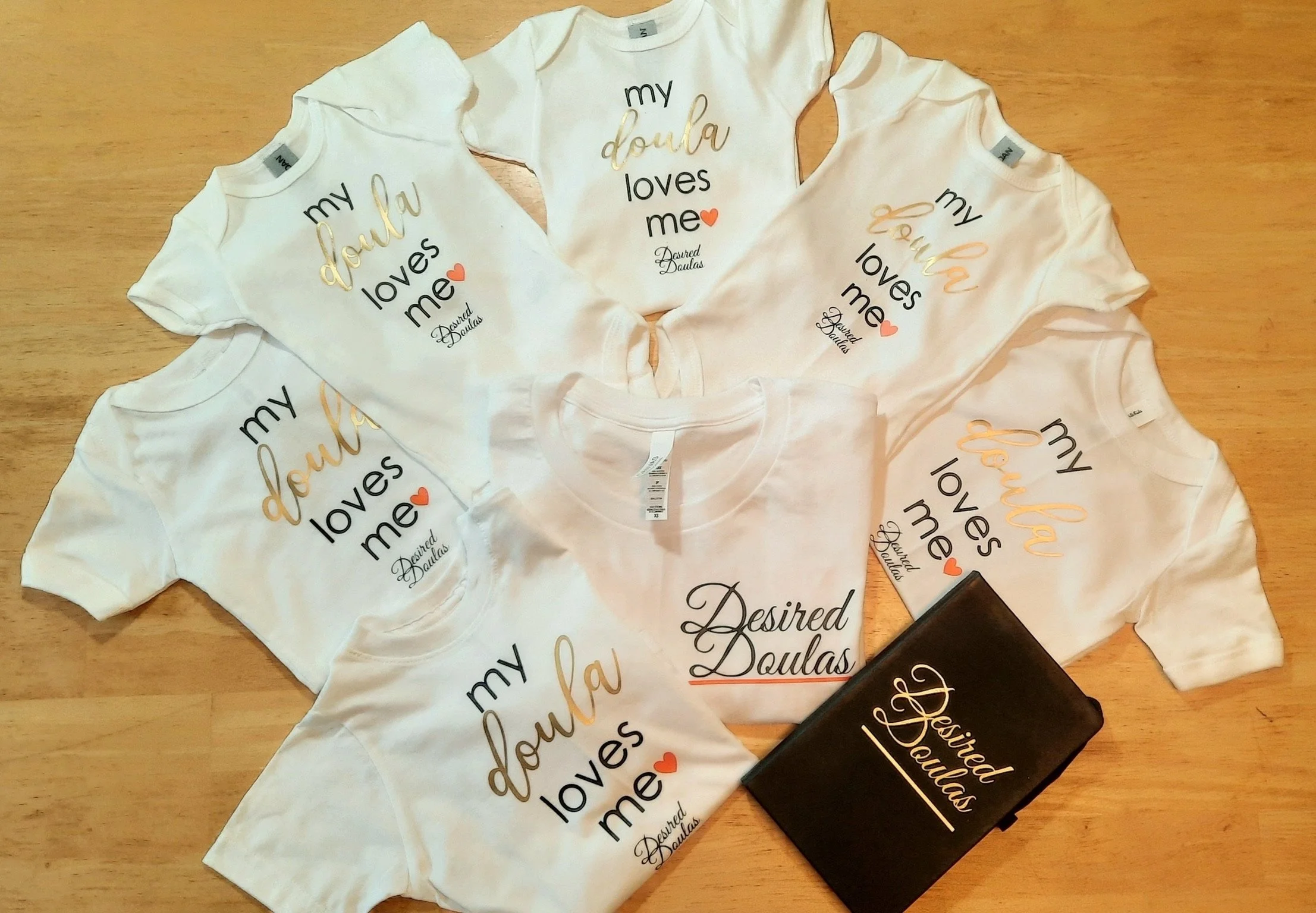 Assorted white baby onesies and a t-shirt with "my doula loves me" and "Desired Doulas" printed on them, alongside a black notebook labeled "Desired Doulas."