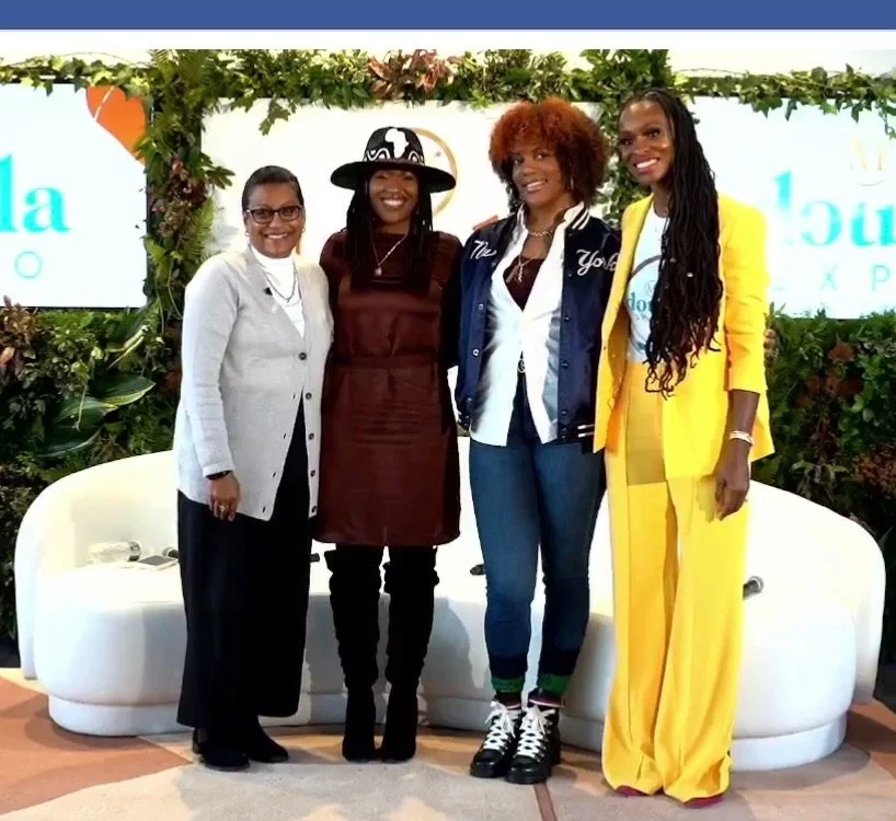 Four women standing together indoors, smiling, in front of a decorated backdrop with foliage and branding. One woman wears a light grey blazer, another a brown dress with a black hat, another a blue jacket with jeans, and the last one a bright yellow