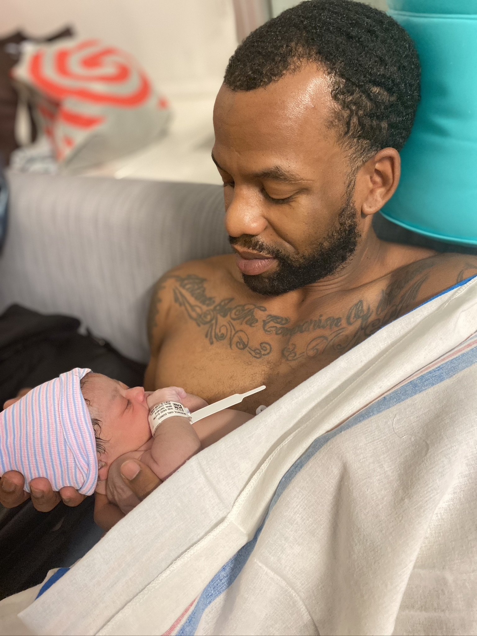 A man holding a newborn baby wrapped in a hospital blanket, with a striped cap on the baby's head. The man is shirtless with tattoos visible and is looking at the baby.