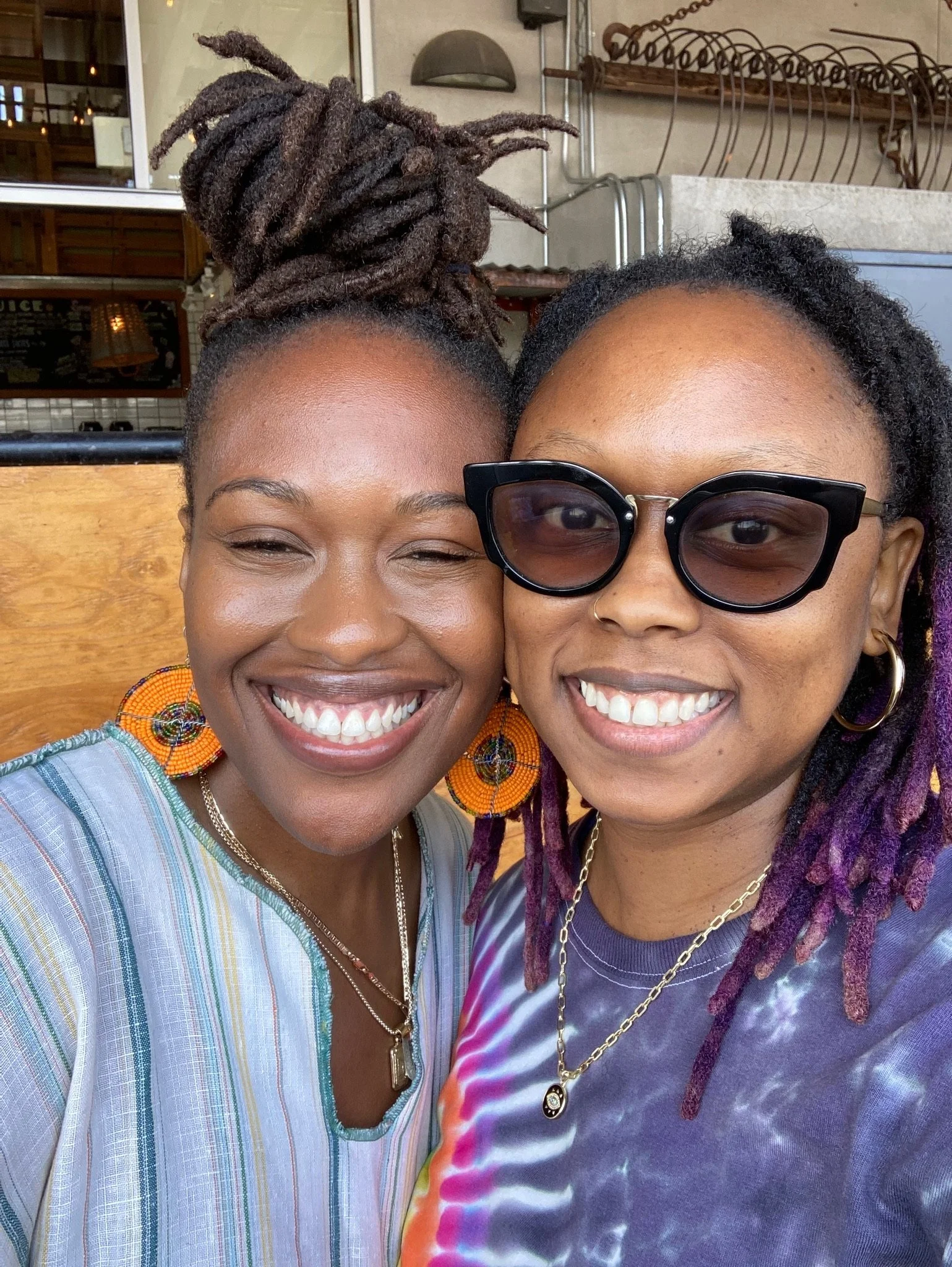 Two people smiling and taking a selfie, one wearing a striped shirt and large orange beaded earrings, and the other wearing sunglasses and a tie-dye shirt, both with braided hair.