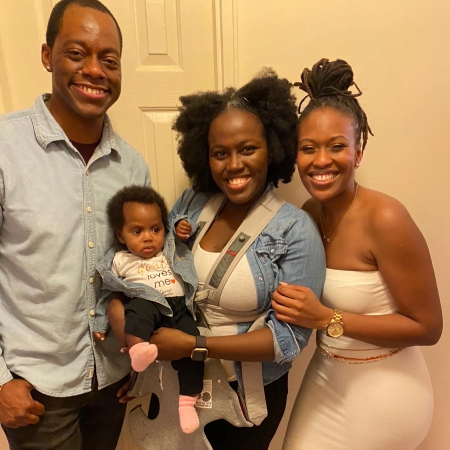 A smiling group of three adults and one baby. The man on the left is wearing a light blue shirt, while the woman in the middle holds a baby wearing a "God loves me" onesie, and another woman on the right is dressed in a white outfit. They appear to b