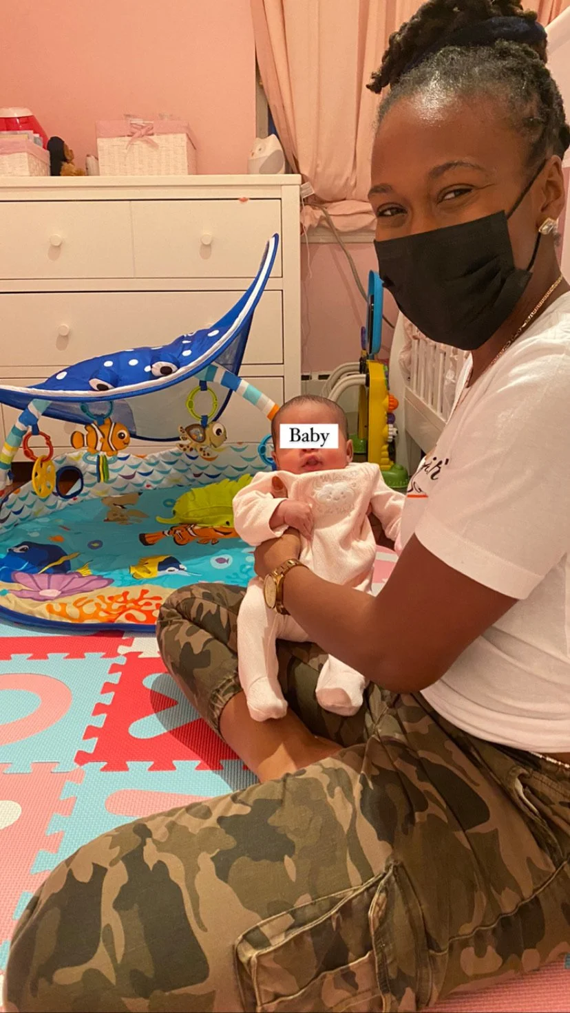 Person wearing a mask holding a baby, sitting on colorful play mat, with toys and a play gym nearby in a nursery.