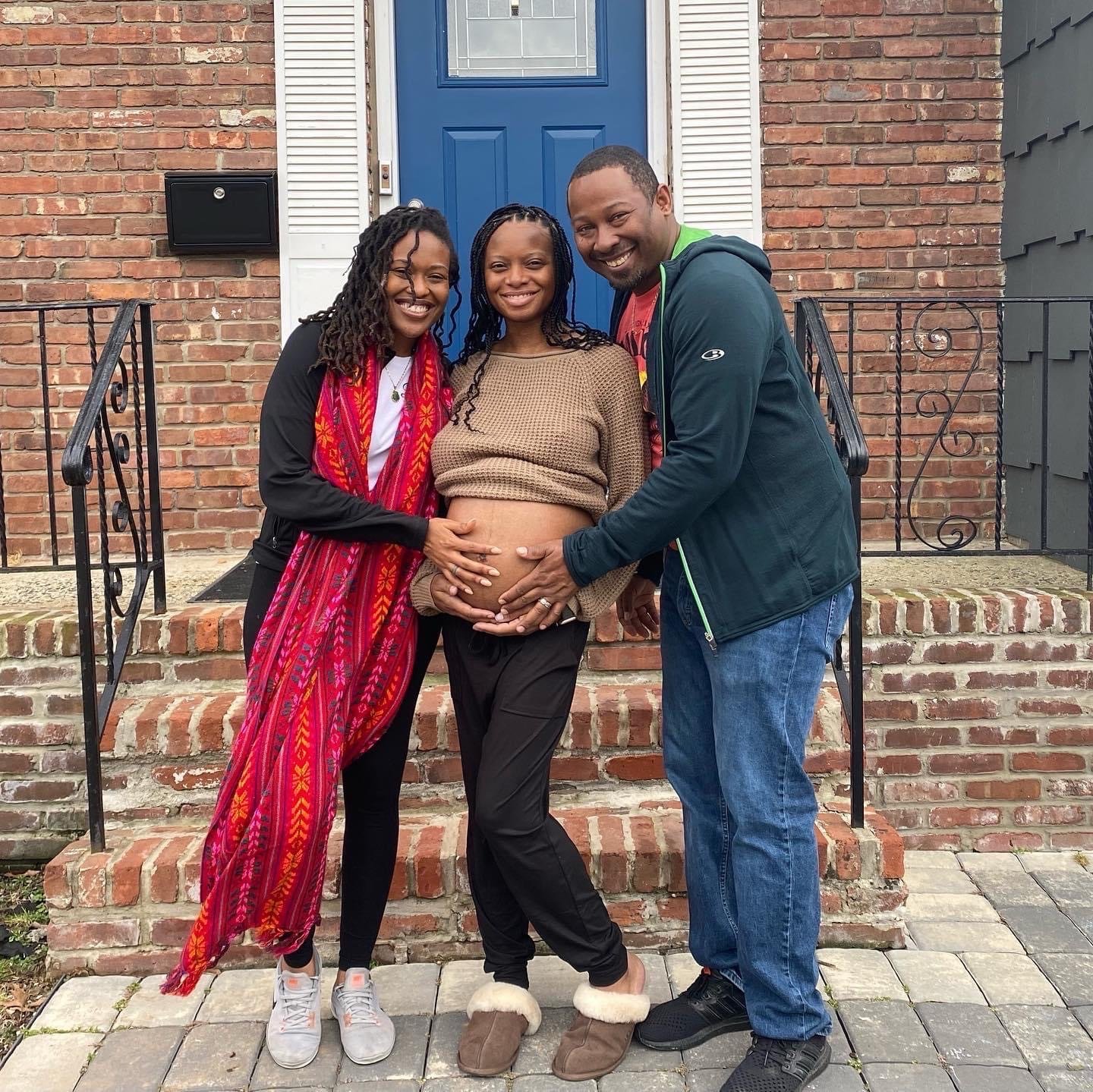 Three people standing in front of a brick house with a blue door. The person in the middle, who is pregnant, is being affectionately embraced by the two individuals on either side. They are all smiling and touching the pregnant belly.