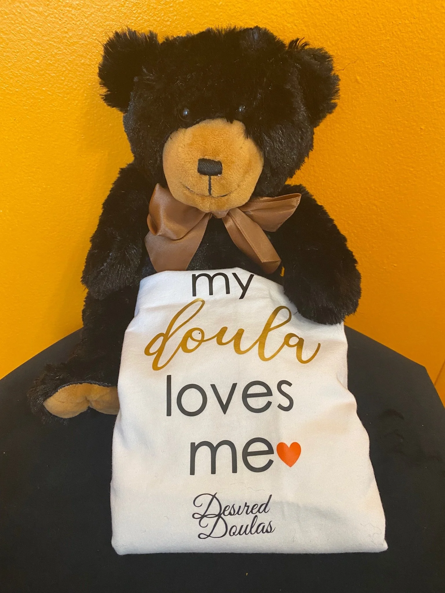 A stuffed black bear with a brown bow holds a white cloth with the text "my doula loves me" and "Desired Doulas" against an orange wall.