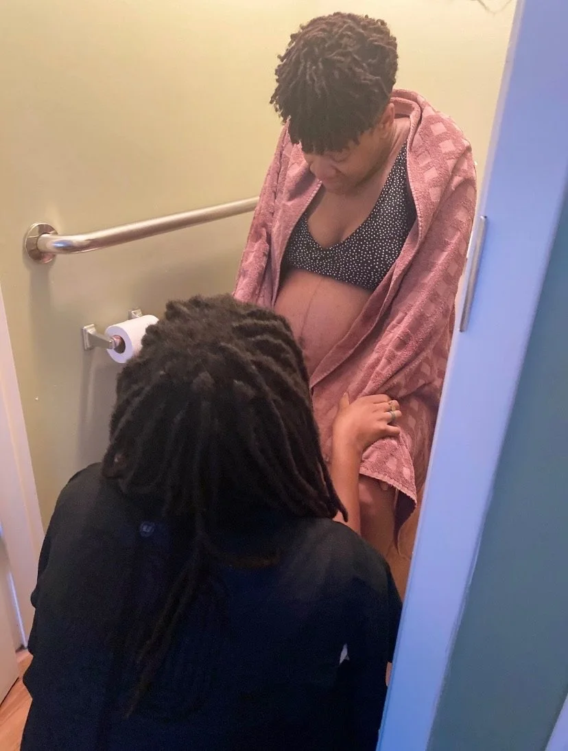 A pregnant woman sitting on a toilet in a bathroom, wearing a black polka dot bra and a pink robe. Another person, crouched down, is offering support, with hands on the pregnant woman's legs.