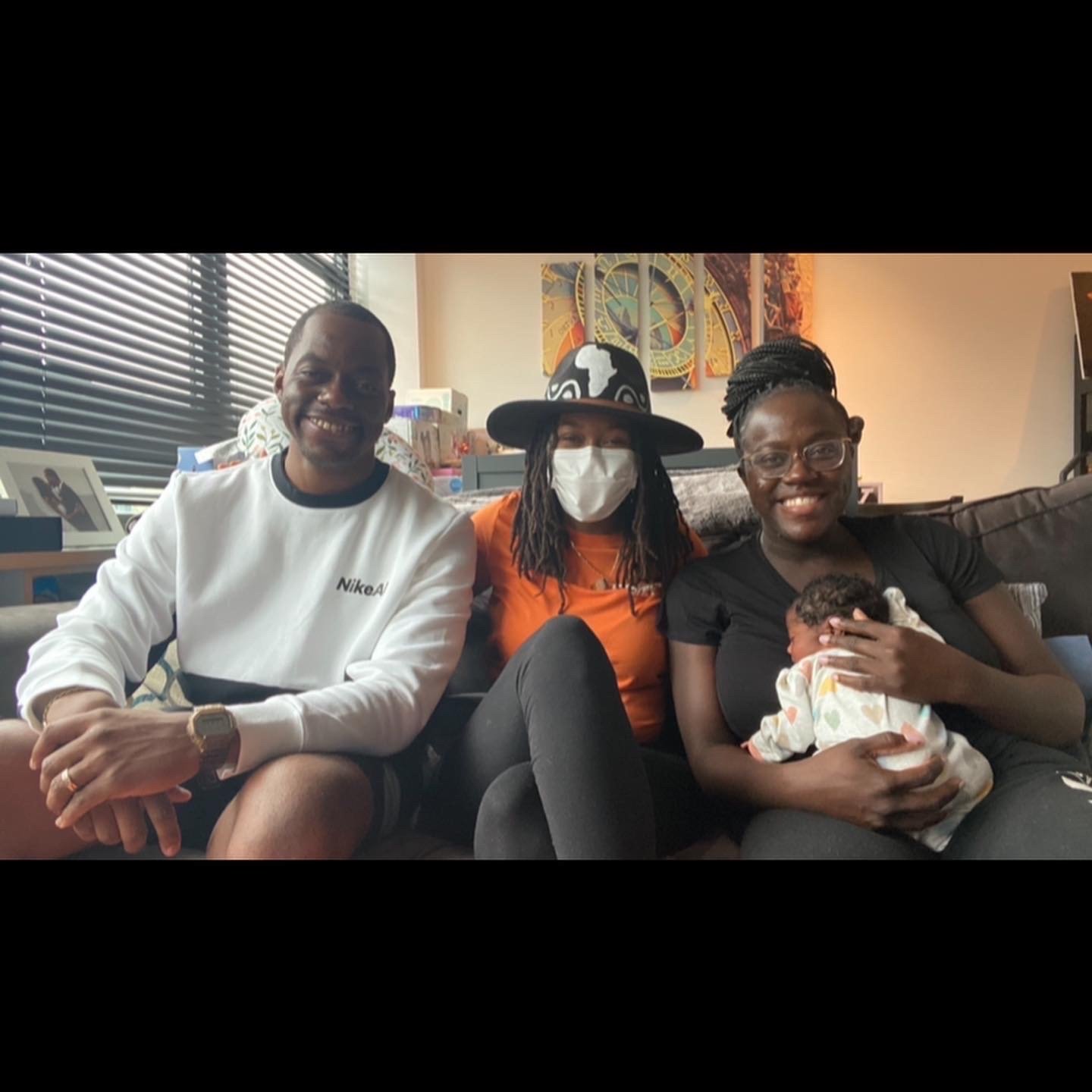 Group of three adults sitting on a couch with a baby, one adult wearing a mask and hat, smiling in a cozy living room.
